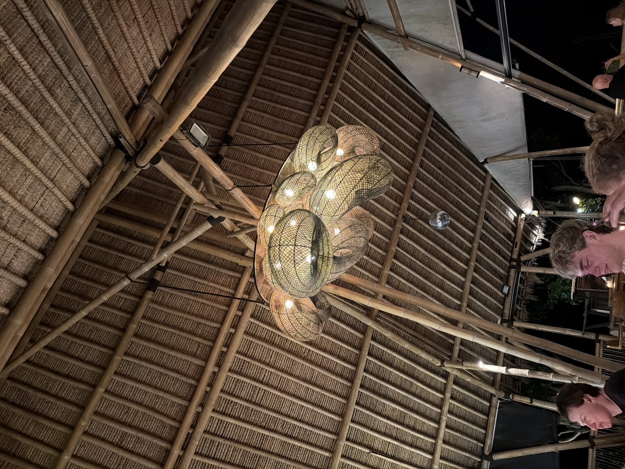 The image displays an upward perspective of the interior ceiling of a building, likely a restaurant or bar, at night in Nusa Penida, Indonesia. The dominant architectural elements consist of a high, vaulted ceiling constructed from a network of sturdy bamboo poles serving as structural beams and rafters. The roof itself is made of tightly woven thatch or natural fibers, creating a textured pattern across the ceiling.
A prominent, multi-component chandelier hangs centrally, featuring numerous spherical and ellipsoid lamps encased in intricate, woven, net-like shades. These lamps emit a warm, glowing light that illuminates the space. Additional lighting includes a rectangular spotlight mounted on a bamboo beam and a small, reflective disco ball hanging towards the left. On the upper right, a section of the roof structure is covered with white fabric or canvas.
Along the right edge of the frame, the partial profiles of at least three individuals are visible: two men and one woman. They appear to be seated or standing, looking downwards, suggesting they are engaged in conversation or activities at a table out of view. The overall ambiance is one of a relaxed, tropical evening social setting. There is no visible text in the image.