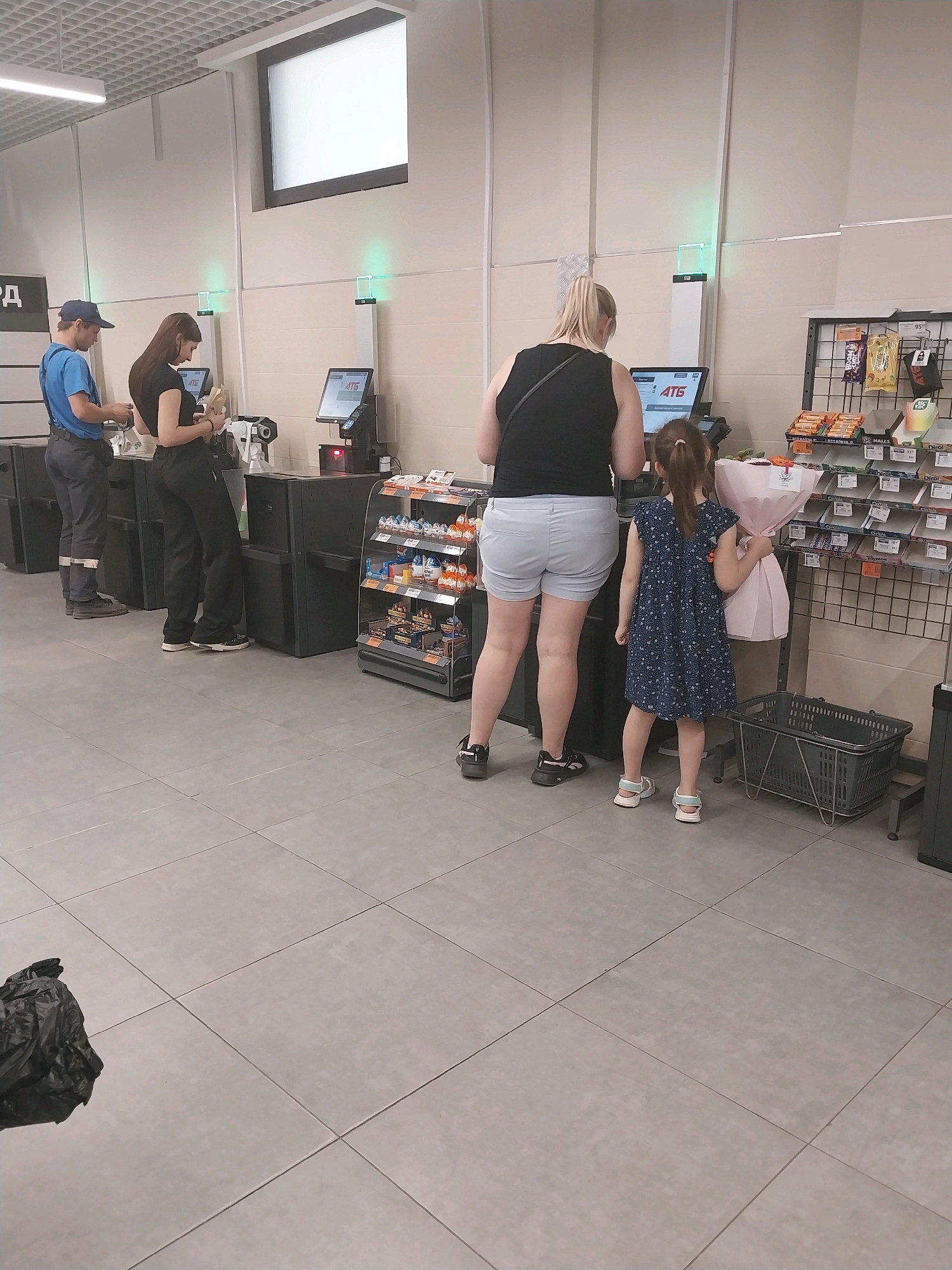 In an indoor retail environment, possibly a supermarket, four individuals are visible. On the left, a male wearing a blue shirt and cap stands at a checkout counter, interacting with a digital screen. To his right, a female with long dark hair, dressed in black, also stands at a counter, holding a small item. Further to the right, a female with blonde hair pulled back in a ponytail, wearing a black top and light shorts, is positioned at another counter, facing a screen. A young female, wearing a blue patterned dress and sandals, stands beside the blonde-haired woman, holding a bouquet of flowers wrapped in pink paper. The environment features tiled flooring and displays of goods on shelves behind the individuals. The scene appears to be at a self-checkout area in a store.