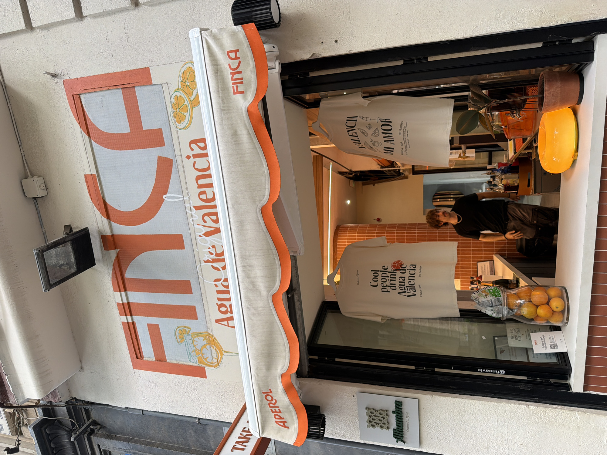 A storefront with an awning displays "FINCA AGUA de VALENCIA" in large orange lettering. Underneath the awning, two t-shirts are displayed. One t-shirt reads "Cool people drink Agua de Valencia" and the other reads "VALENCIA MI AMOR". A man wearing a black t-shirt and brown pants stands behind a counter, serving customers. A glass jar filled with oranges sits on the counter. The storefront appears to be a bar or cafe. The location is unknown.