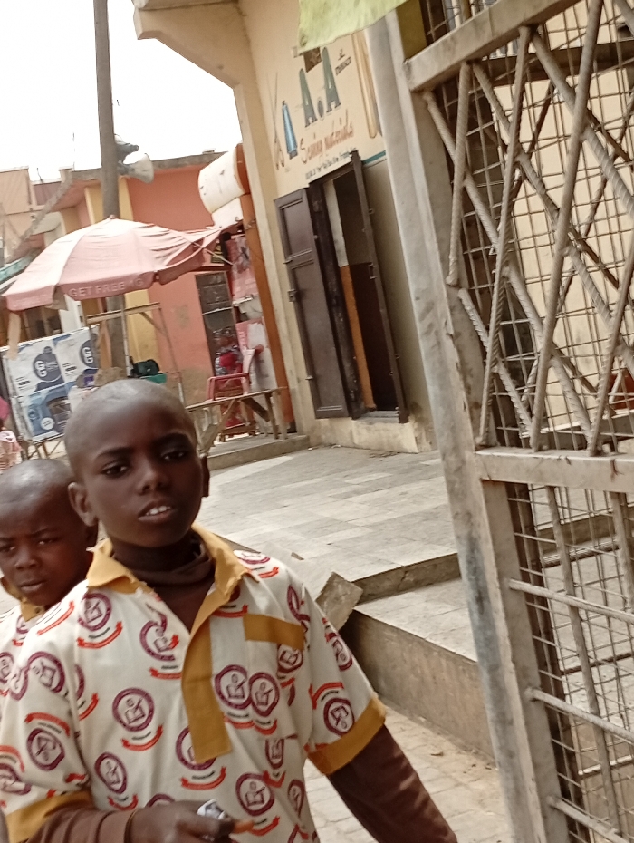 In Gawuna, Nigeria, two young males are captured outdoors in what appears to be a commercial street. The foreground shows a male with a shaved head and dark complexion, wearing a white short-sleeved shirt patterned with reddish-brown circular motifs and featuring mustard yellow collar and sleeve cuffs. A dark fabric is visible around his neck, and he holds a small, light-colored object in his left hand. Partially obscured behind and to the left of him is a second male, also with a shaved head. They stand on a grey paved surface, with concrete steps visible behind them. To the right, a weathered metal gate with a diamond lattice pattern partially frames the scene. In the background, a light-colored building displays a sign with "A.A Sewing materials" and illustrations of scissors and thread. Its dark brown double doors are partially open. Further to the left, a red market umbrella with "GET FREE" text and various items are visible, suggesting a street vendor's stall.