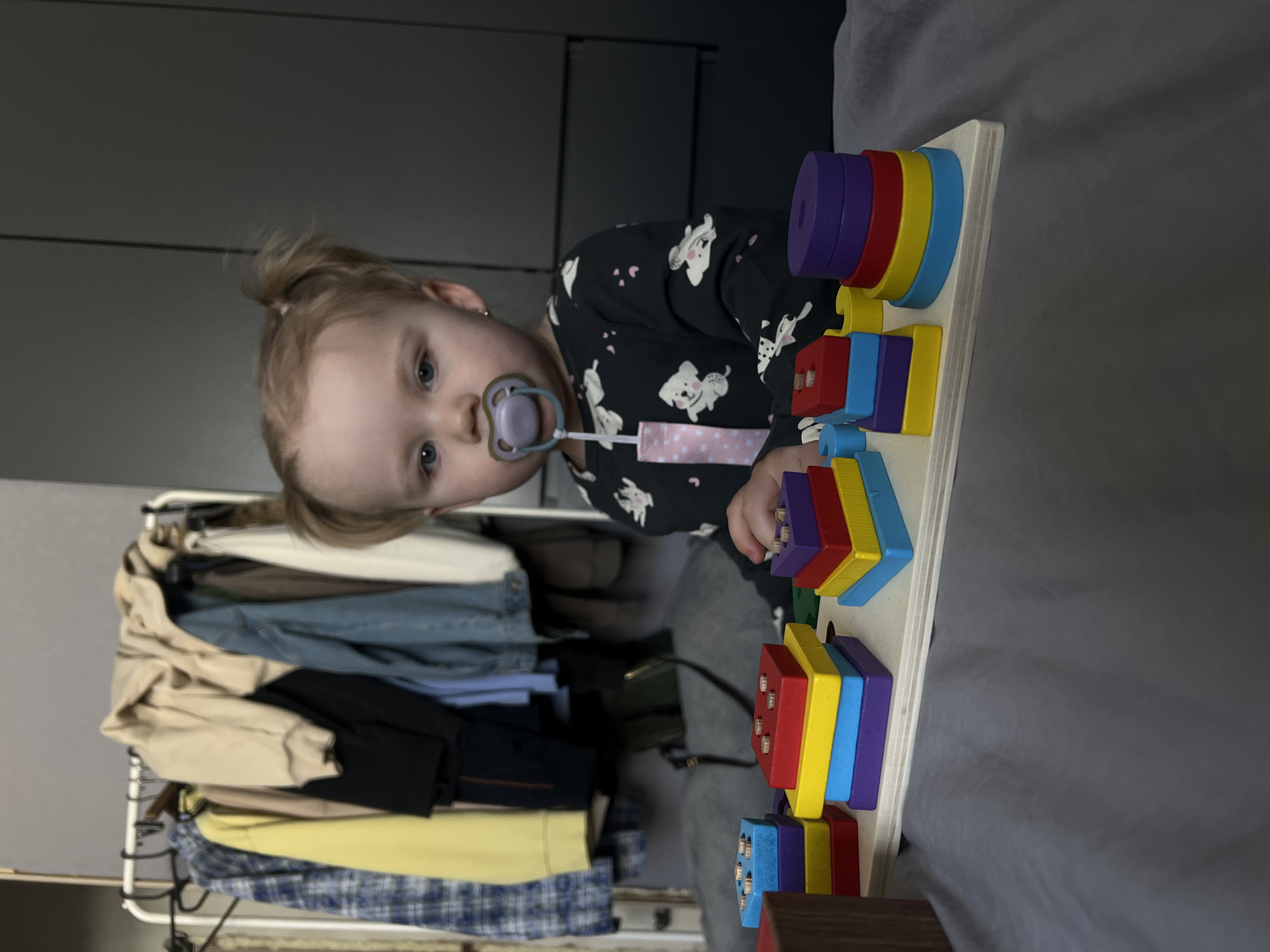 An indoor scene in the city of Leninakan, Russia, depicts a young child seated on a dark gray fabric surface, likely a bed. The child, with fair skin, blue eyes, and light hair styled in two small pigtails, looks directly forward. A purple pacifier is in her mouth, attached by a pink and white patterned strap. She wears a dark long-sleeved garment adorned with white dog figures and pink paw prints. In the foreground, a wooden educational toy board featuring multiple colorful geometric stacking shapes (red, yellow, blue, purple squares, rectangles, and circles) rests on the dark gray surface. The child's left hand is partially visible near one of the toy's stacks. The background to the left displays a white metal garment rack laden with assorted clothing, including a blue plaid shirt, a yellow sweatshirt, a beige hoodie, and a denim jacket. A light gray wall is visible behind the rack. To the right, a dark gray furniture piece, possibly a wardrobe or headboard, extends upward.