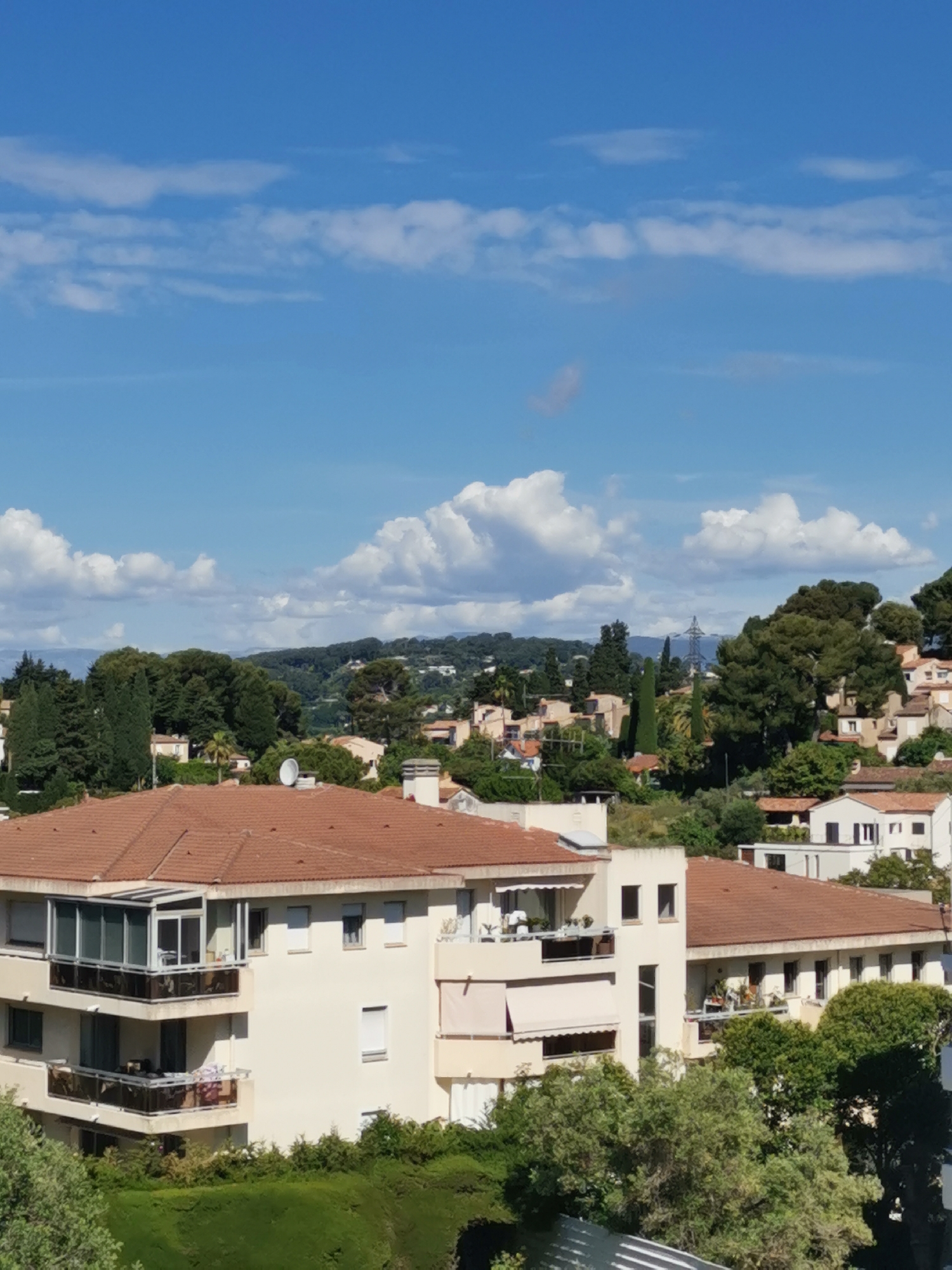 The image captures a bright, sunny day in an urban area, likely a residential district. The sky is predominantly blue with scattered, puffy white clouds. The foreground is dominated by buildings with terra-cotta tile roofs and light-colored facades, indicating a possible Mediterranean architectural style. Trees and greenery are interspersed amongst the buildings, creating a somewhat lush environment. In the mid-ground, the cityscape extends further with more buildings, trees, and a distant hillside or mountain range. An electrical power tower is visible among the trees in the mid-ground. The image appears to have been taken from an elevated position, providing a wide, clear view of the city and surrounding landscape.