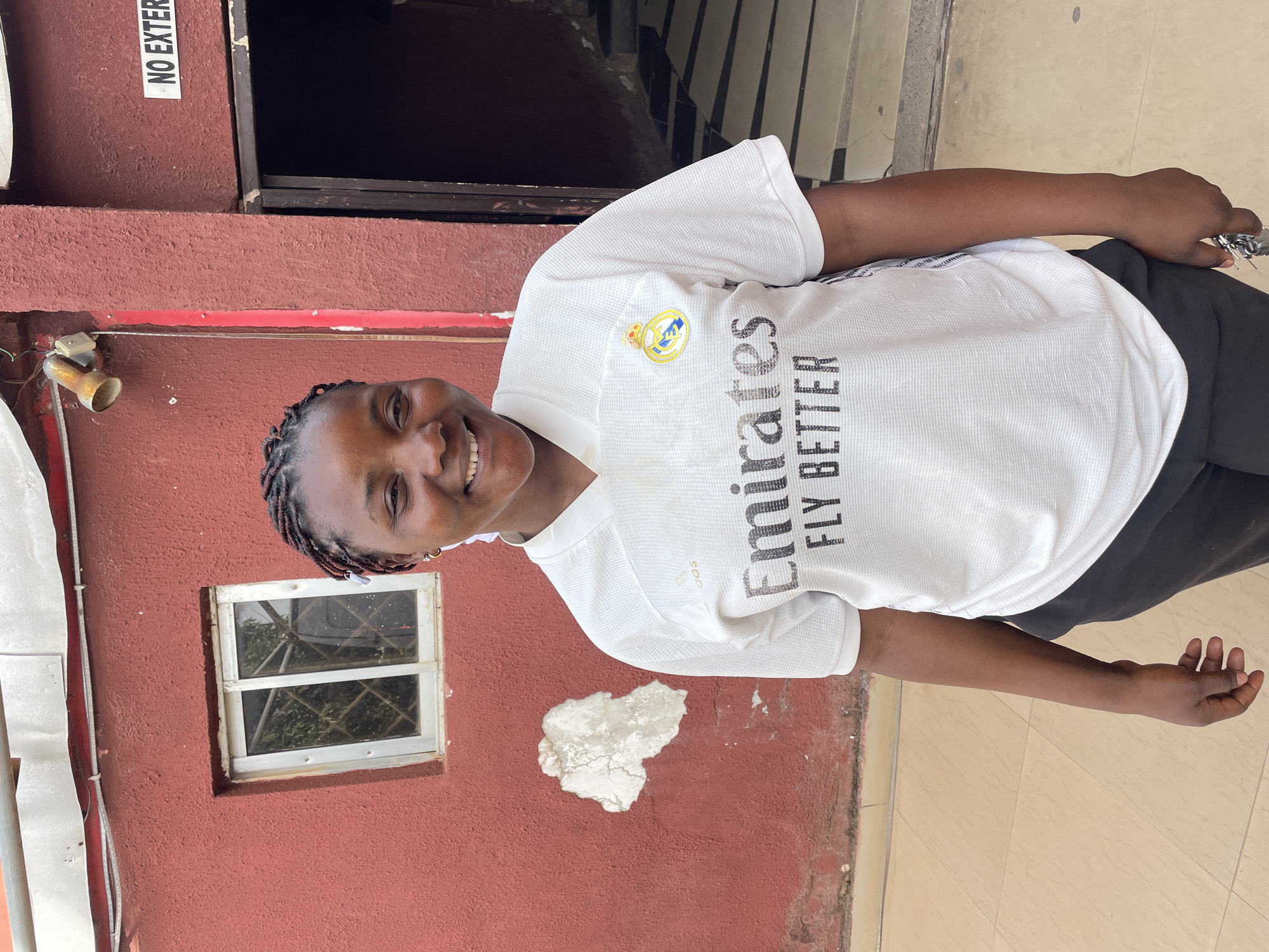 A young individual with dark skin and braided hair is captured smiling broadly at the camera. They are wearing a white short-sleeved football jersey, which prominently displays a golden Real Madrid crest on the left chest and the text "Emirates FLY BETTER" in black across the front. Dark trousers or shorts complete their attire.
The background is dominated by a textured, reddish-brown building wall. To the left of the person, a small, square window with white frames and a security grill is visible, offering a glimpse of green foliage outside. Above the person's head, a dark, recessed doorway or opening leads further into the building. A patch of white plaster stands out against the red wall, indicating some wear. Various fixtures, including a metallic bell-shaped object (possibly an old light fitting) and white vertical conduits, are attached to the wall. The ground beneath the person is a light-colored tiled surface.
The image was taken during the daytime, indicated by the natural and even lighting. The subject's wide and joyful smile conveys a positive and friendly disposition. A white sign with the partially visible text "NO EXTER" (likely "NO EXTERNAL" or "NO ENTRY") is affixed to the upper left part of the wall. The scene is set in an outdoor or semi-outdoor area, characteristic of a building exterior in Jos, Nigeria.