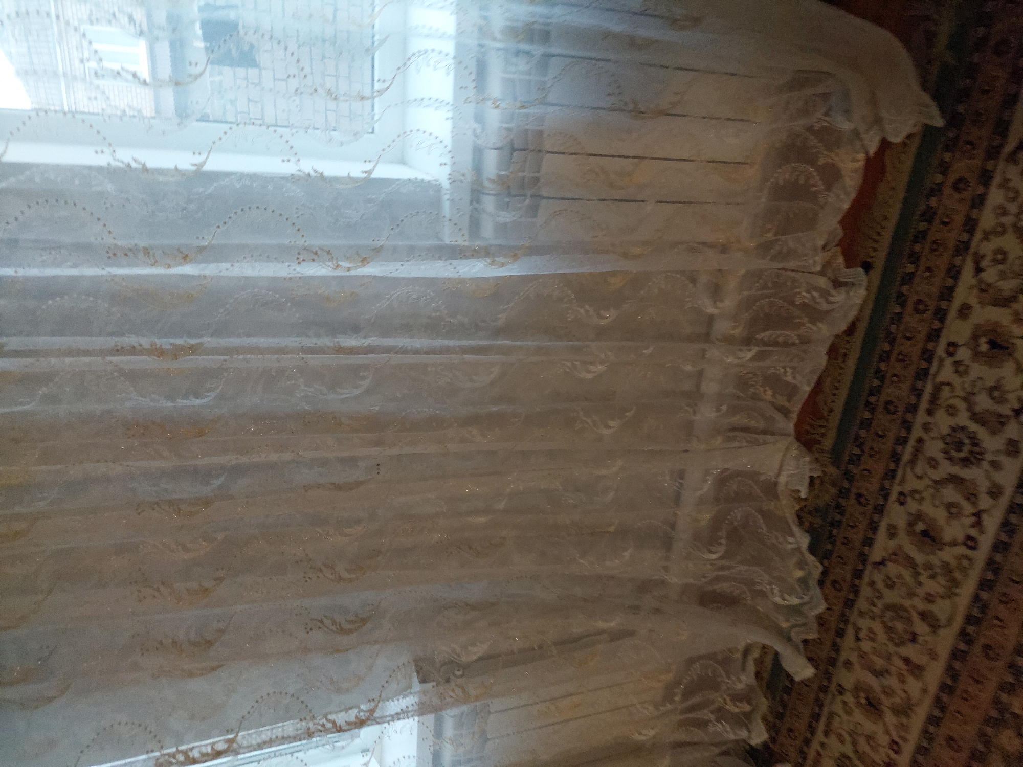 The image shows a close-up of a sheer, white curtain with a gold embroidered pattern of swirling vines and dots. The curtain is hanging in front of a window, partially obscuring a white radiator and a brick building with windows visible outside. A patterned carpet with red and brown tones covers the floor beneath the curtain. The overall impression is of an interior domestic setting, likely a living room or bedroom, in a building located in Makhachkala, Russia.