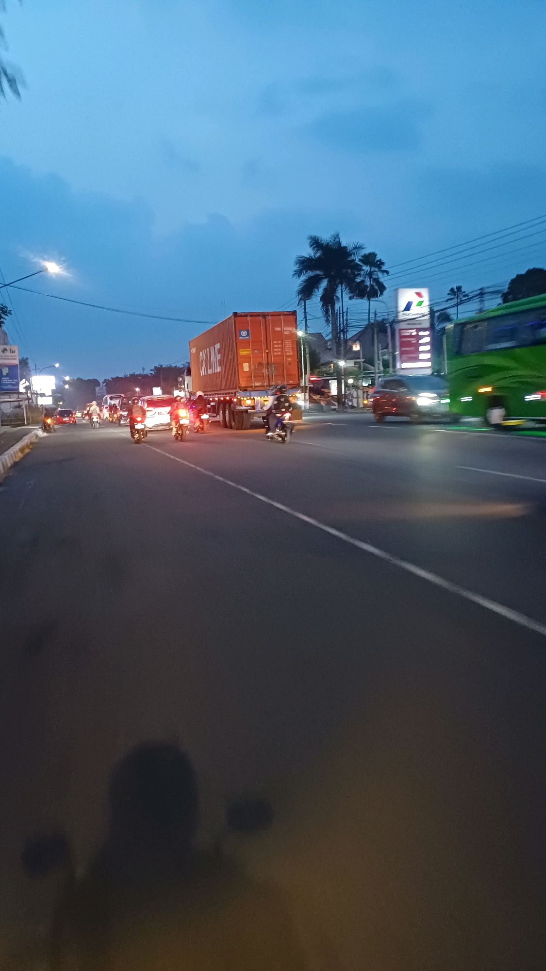 The image depicts a street scene in Kowangan, Indonesia, during twilight. A large orange shipping container truck is prominently featured, stopped on the right side of the road. Several motorcycles are also present, with some riders visible. On the far right, a green bus is partially visible. To the left of the truck, traffic lights and streetlights illuminate the road. Palm trees and other vegetation are scattered throughout the background. The sky is a deep blue, indicating the late hour. The road surface is asphalt, with a white line marking the lanes. The overall impression is of a busy urban environment at the end of the day.