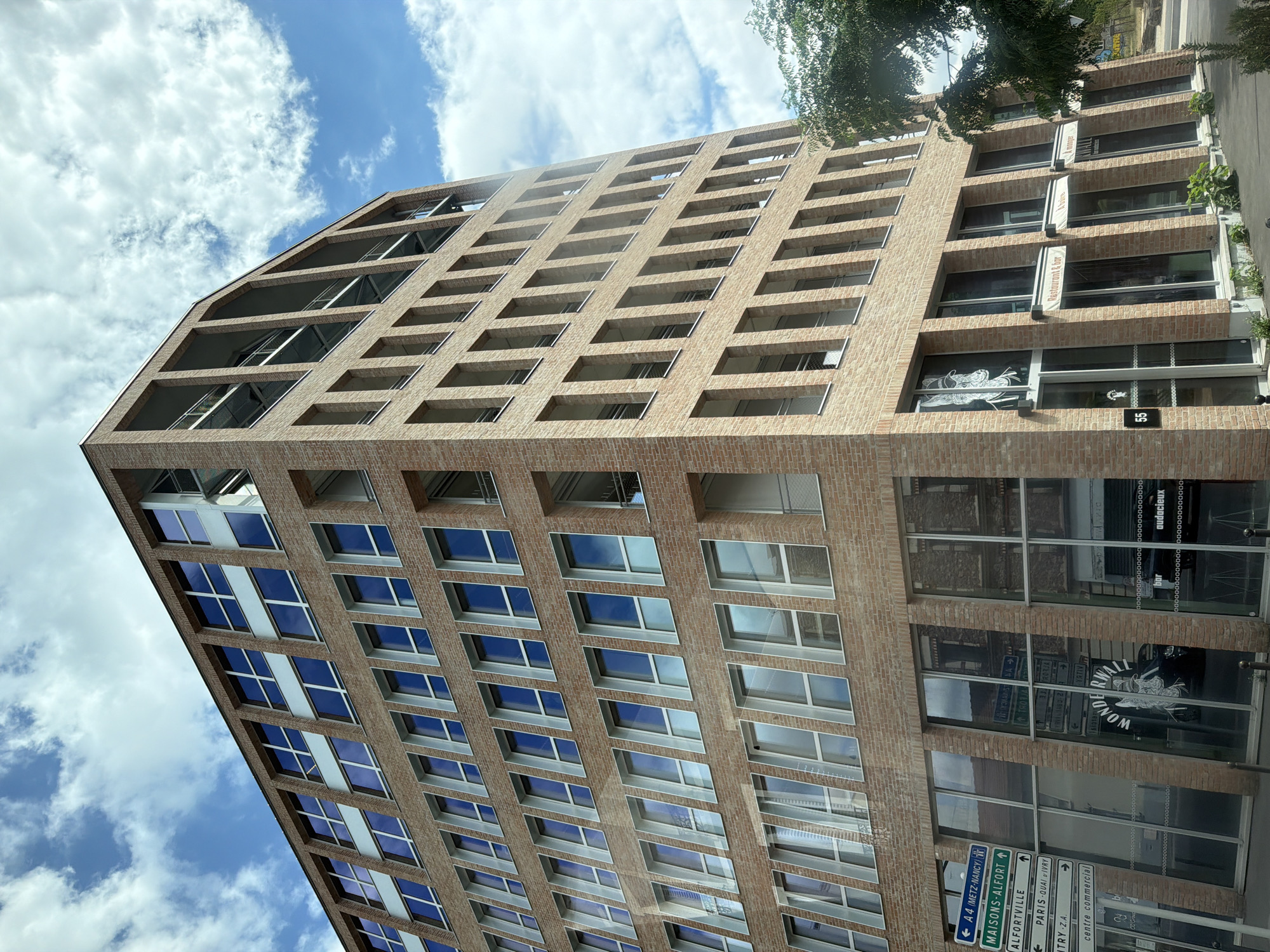This media file shows a modern brick building with many windows, seen from a low angle looking upwards. The building has a unique, angular design, with recessed balconies or windows creating a grid-like pattern. The sky is visible above, with white clouds against a blue background, suggesting it is daytime. A tree with green leaves is partially visible at the top right, adding a touch of nature to the urban scene.
On the ground floor of the building, there are large glass windows with some reflective advertisements and signage. Visible text includes "Restaurant & bar," "55," and what appears to be a stylized drawing with the word "WONDERWALL." Further down the street, street signs are visible, with text in French indicating directions to "MAISONS-ALFORT," "ALFORTVILLE," "PARIS QUAI D'IVRY," and "centre commercial." These signs provide strong location cues for Ivry-sur-Seine, France.
The overall impression is that of a contemporary urban setting, possibly a commercial or residential building, with clear signage indicating businesses and local destinations. The weather appears to be pleasant, with a mix of sun and clouds. There are no people or specific events depicted, focusing instead on the architecture and urban environment.