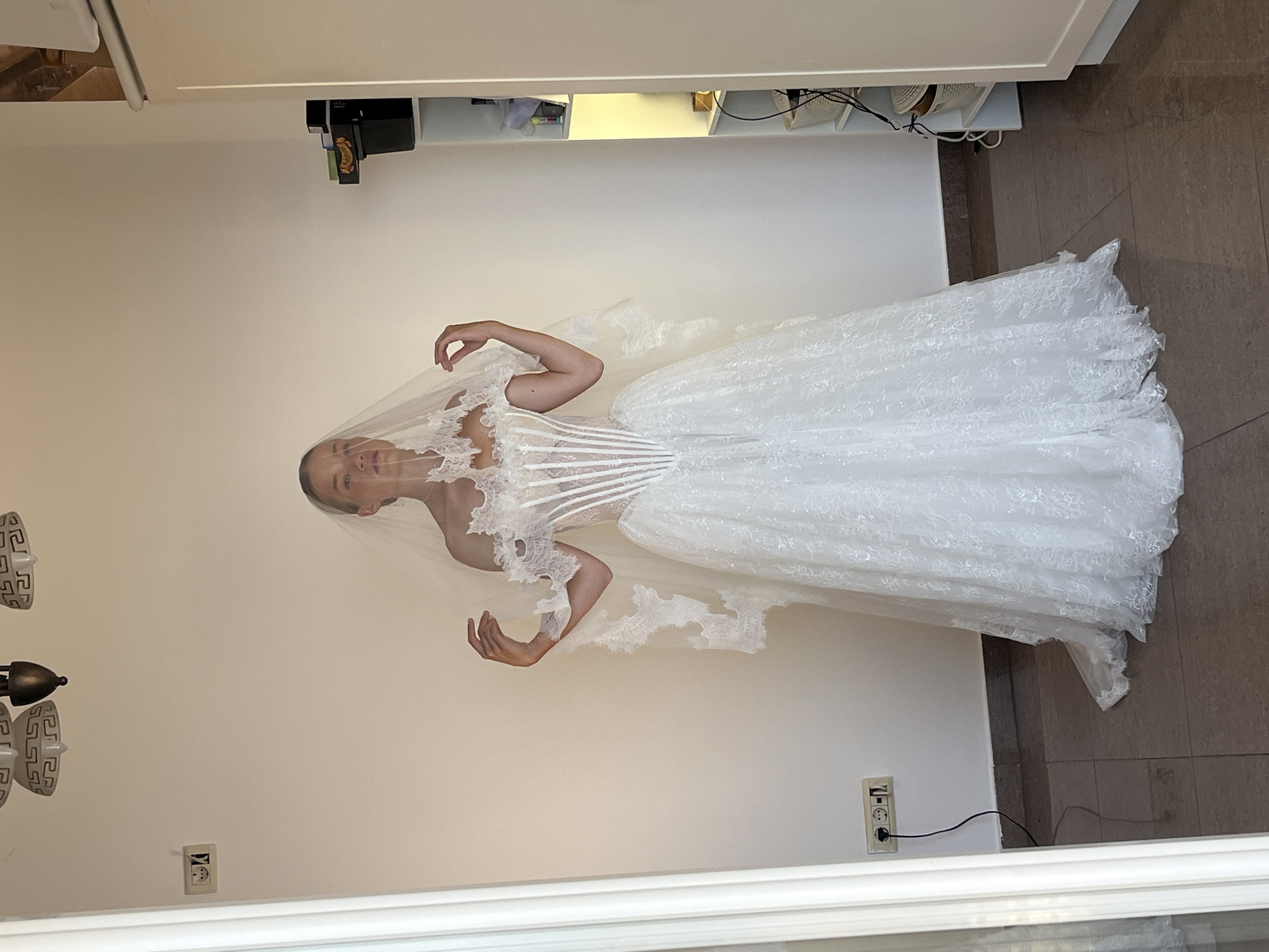 A young woman, wearing a white lace wedding dress and veil, stands facing forward in a room. The dress features a fitted corset bodice with vertical boning and a full, floor-length lace skirt. The veil is also made of lace and drapes over her shoulders and head. The background is a plain, off-white wall with a light-colored tile floor. To the left, a white doorway or frame is visible, and to the right, a white shelving unit holds various items. Two electrical outlets are mounted on the wall to the left of the woman, with a black cord plugged into one. The room appears to be indoors, possibly a residential setting, and given the details, it is highly likely located in Budva, Montenegro.