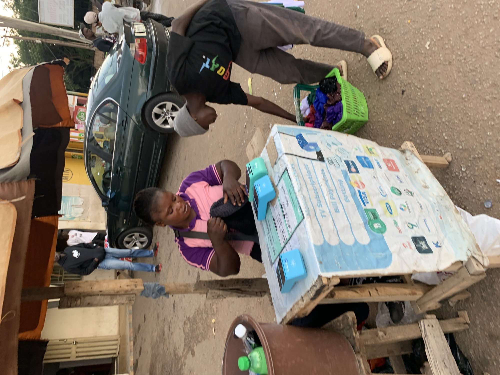 The image depicts an outdoor scene in Jos, Nigeria, likely during the day given the natural lighting. The setting appears to be a street or market area, characterized by a dirt ground and various makeshift structures.
The main subjects are two individuals, an adult male and an adult female, engaged in a transaction. The man, wearing a dark hoodie, grey beanie, and brown pants, is leaning over a table. He is reaching towards the woman, who is seated and wearing a pink and purple striped shirt. She appears to be handling a transaction, possibly involving the two blue electronic devices on the table in front of her. A green plastic basket filled with colorful fabrics is placed near the man.
The table is covered with a white sheet displaying various logos, including VISA, and text that seems to indicate services like "TV Subscription," "Bill Payment," and "Setting." This suggests the table is a point of sale for various services. A dark-colored sedan is parked behind the man, with its rear door open. In the foreground, a large brown plastic container is visible, containing a green bottle and other items.
In the background, another person in a white hoodie is standing near the car. The overall atmosphere seems casual and transactional, typical of a bustling informal market setting. The time of day appears to be late afternoon or early evening, with the soft light suggesting the sun is not high in the sky.