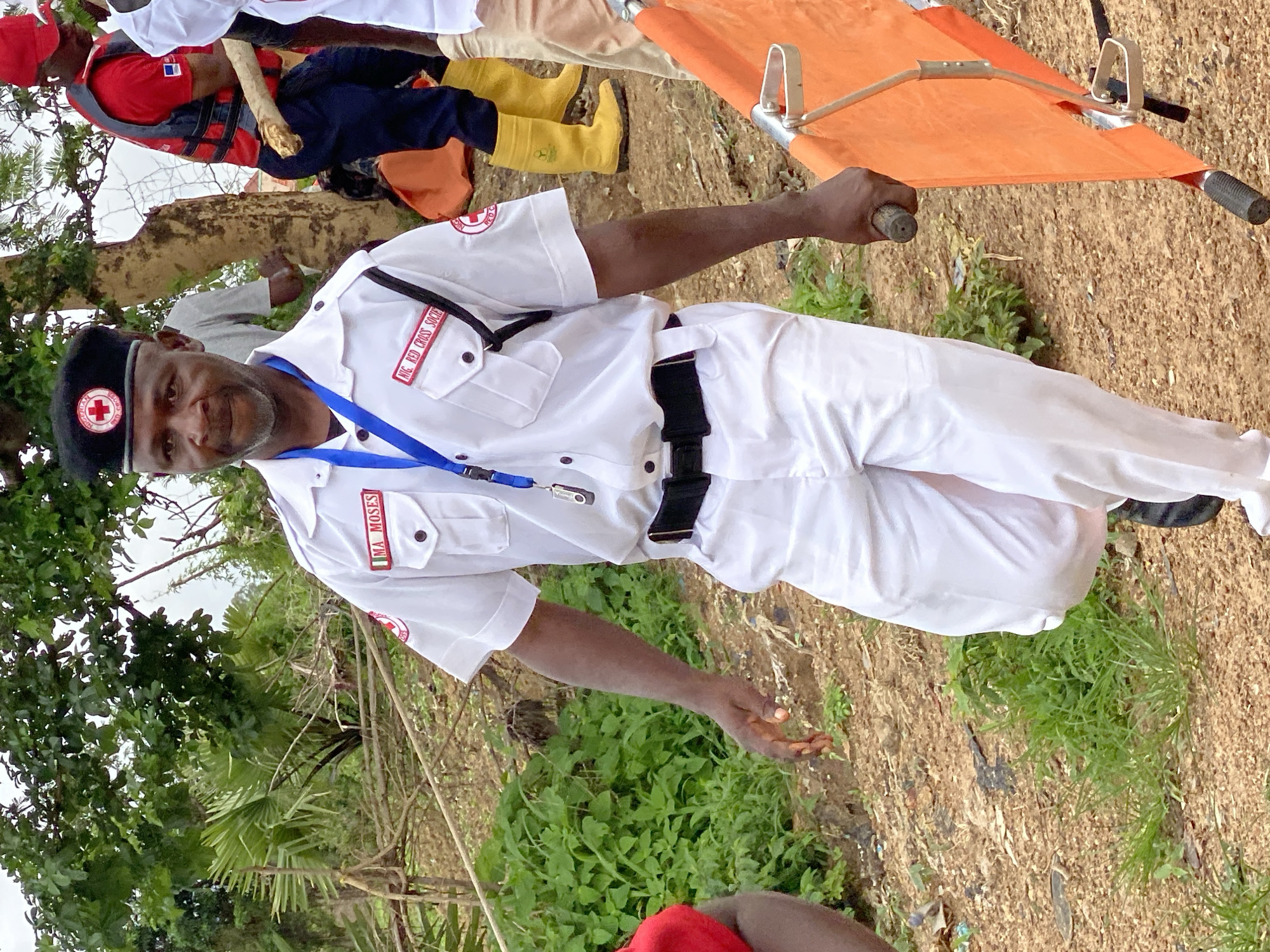 In Kawo, Nigeria, a man wearing a white uniform with red trim and a black beret with a red cross emblem stands outdoors. He has a name tag on his uniform that reads "IMA MOSES." A blue lanyard with a badge is around his neck. He is holding the handle of an orange stretcher. Behind him, another person in a red vest and dark pants, also wearing a red cross emblem, is visible. The ground is covered in dirt and vegetation, and trees are in the background. The scene appears to be during the daytime under an overcast sky.