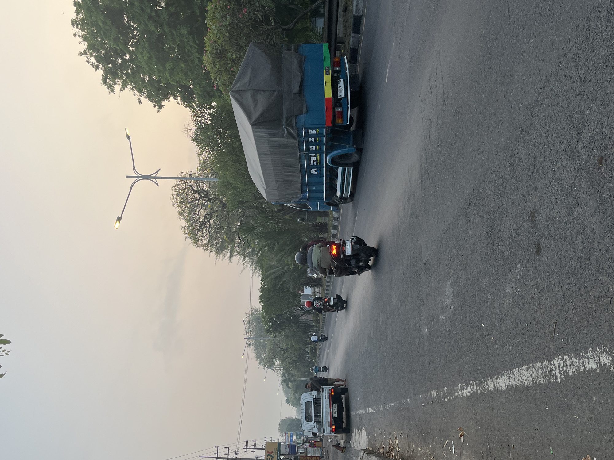 In Kludan, Indonesia, a blue truck with a tarp-covered bed is driving on a paved road. The truck has license plate number N 8069 ND. Several motorcycles are also on the road, with riders wearing helmets and casual clothing. One motorcycle, in the foreground on the right, has two riders. A white truck is parked on the left side of the road, and two people are standing near it. The scene takes place on a street with trees and buildings lining the sides. The sky is hazy with a hint of sunset colors. Streetlights are visible above the trees. The road has white lane markings and appears to be in good condition.