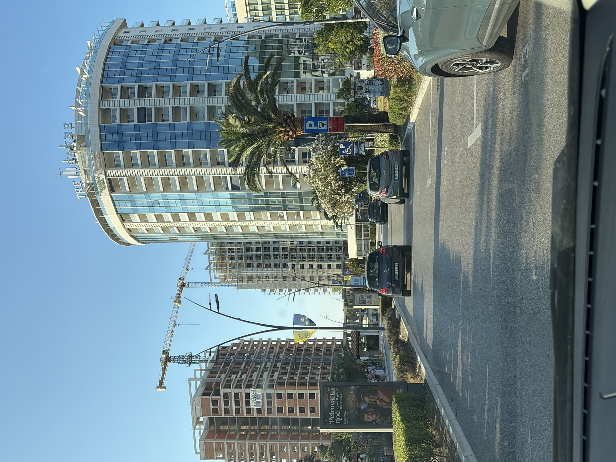 The image depicts a street scene in Budva, Montenegro, with ongoing construction of several buildings. In the foreground, a light-colored SUV is parked in a marked parking space. Further down the road, several cars are driving or stopped. On the right side of the road, a tall, modern building with a curved facade and blue glass windows is visible, with the name "TRE CANNE" prominently displayed at its top. To the left of this building, a palm tree stands tall, accompanied by flowering bushes. Beyond the palm tree, construction cranes are visible, suggesting active building sites. Several other buildings, in various stages of construction, are also present. A billboard with an advertisement for "Petrovačka noć" is situated on the left side of the road, near a patch of manicured hedges. The sky is clear and blue, indicating a sunny day.