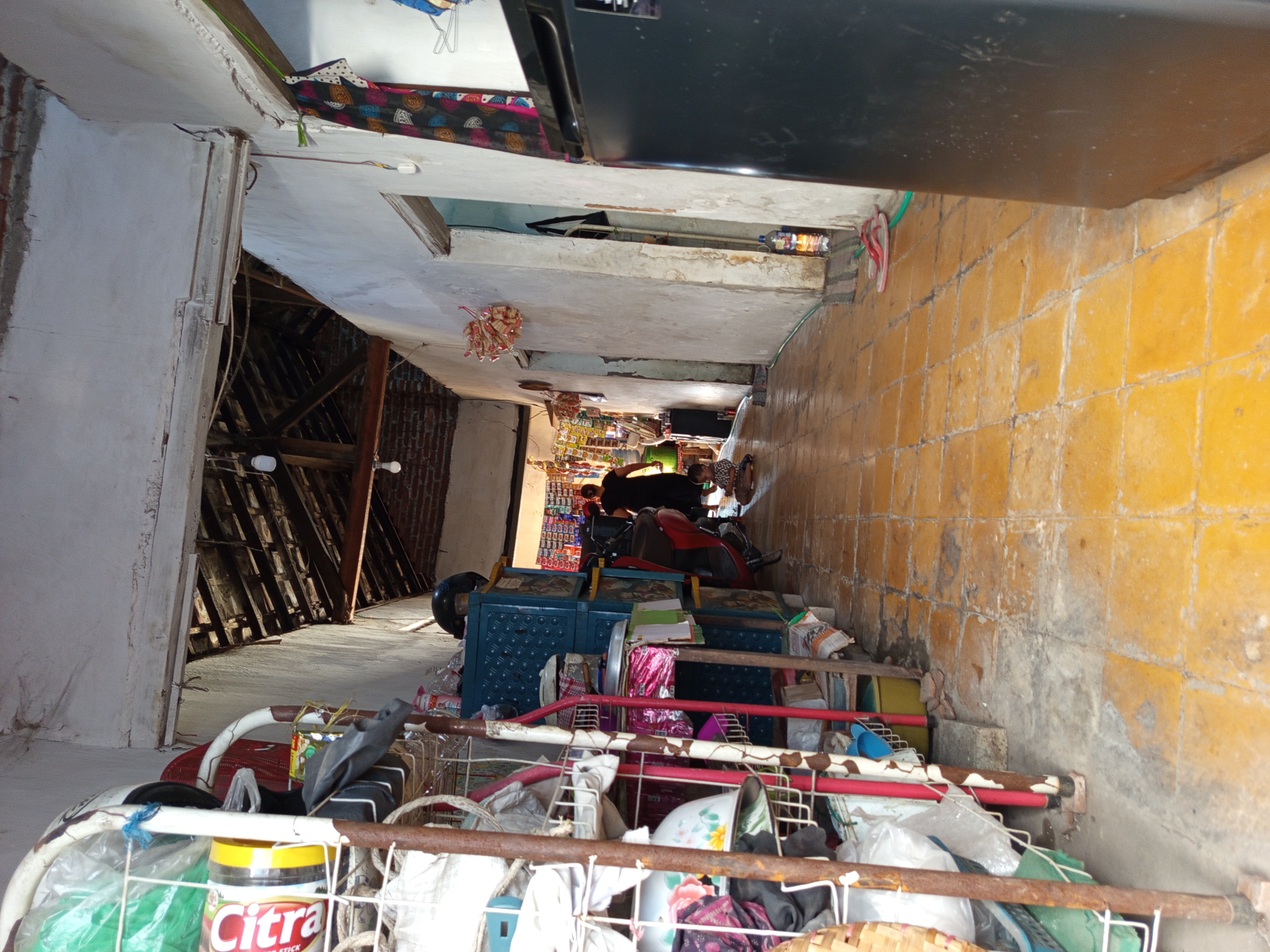 This is a full, eye-level, slightly angled, outdoor shot of a narrow walkway or alley in Tebel, Indonesia. The walkway is paved with yellow square tiles that are dirty and worn. On the left side of the walkway are shelves filled with various items, including plastic-wrapped goods, buckets, and what appear to be cleaning supplies. The shelves are made of white metal. Further down the walkway, to the left, is a red motorcycle and a blue storage unit.
In the distance, at the end of the walkway, is an open area that appears to be a small shop or market, with shelves of goods visible. A woman in a dark dress is walking away from the camera towards this shop. A young child, wearing a light-colored t-shirt and shorts, is sitting on the ground near the middle of the walkway, facing away from the camera.
The buildings on either side of the walkway have white walls and exposed wooden beams and roofing in some areas, suggesting an older or unfinished construction style. Several light bulbs are visible hanging from the ceiling. On the right side of the walkway, closer to the foreground, is a large, black refrigerator. Pink flip-flops are on the ground near the refrigerator. The overall impression is of a busy, utilitarian space within a residential or commercial area.