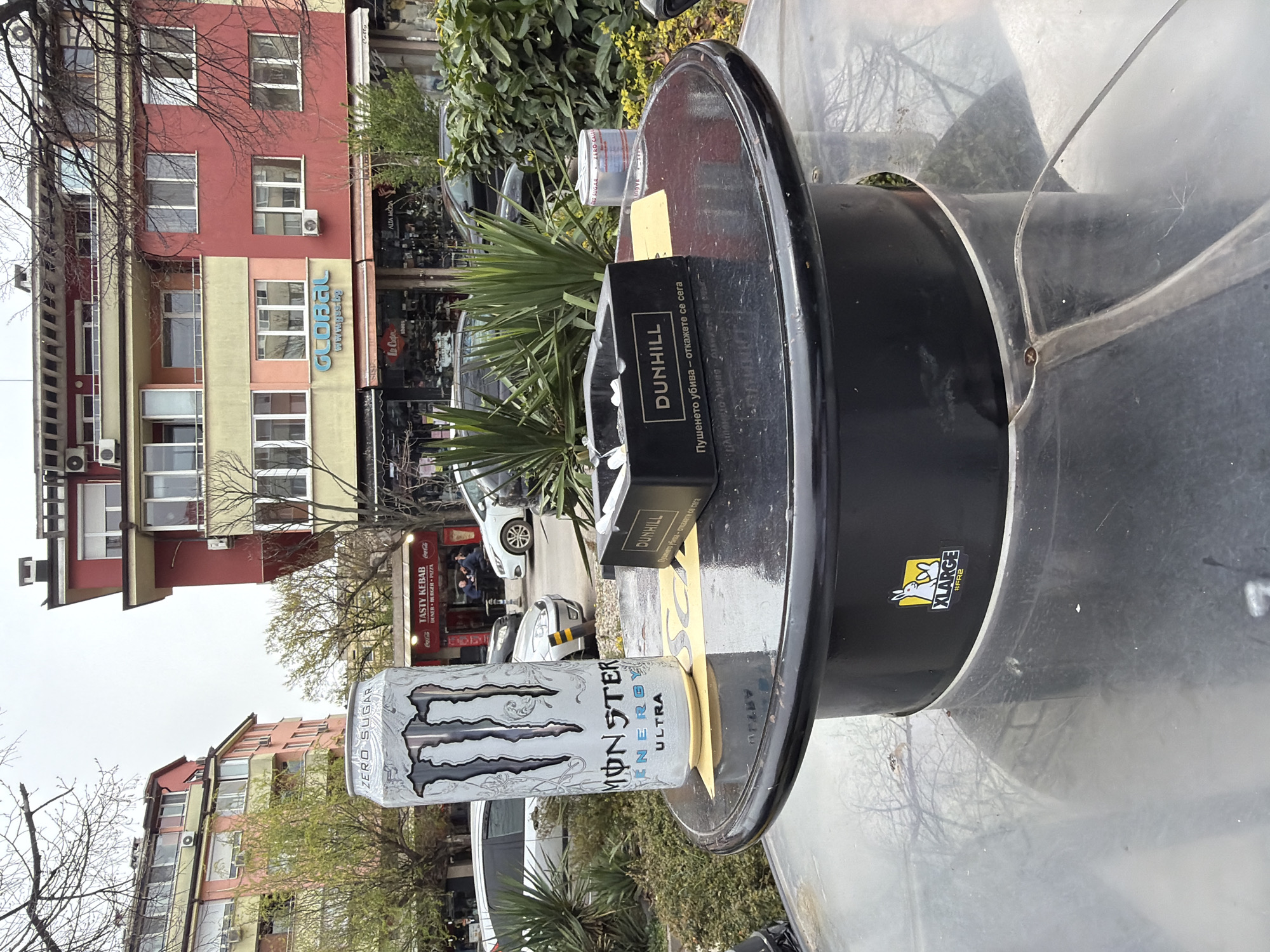 The image depicts an outdoor scene in Sofia, Bulgaria. In the foreground, a black, round-topped trash receptacle is the primary focus, covered in a large plastic cover. A can of Monster Energy Ultra, a Dunhill cigarette pack, and a small plastic container sit on top of the receptacle. The brand sticker of XLARGE #FR2 is also visible on the bin.
In the background, buildings line the street, including one with a sign that says "GLOBAL www.gs.bg". A store with the name "TASTY KEBAB" and "Coca-Cola" signs is also identifiable. The environment consists of urban greenery and parked cars. The sky overhead appears overcast.