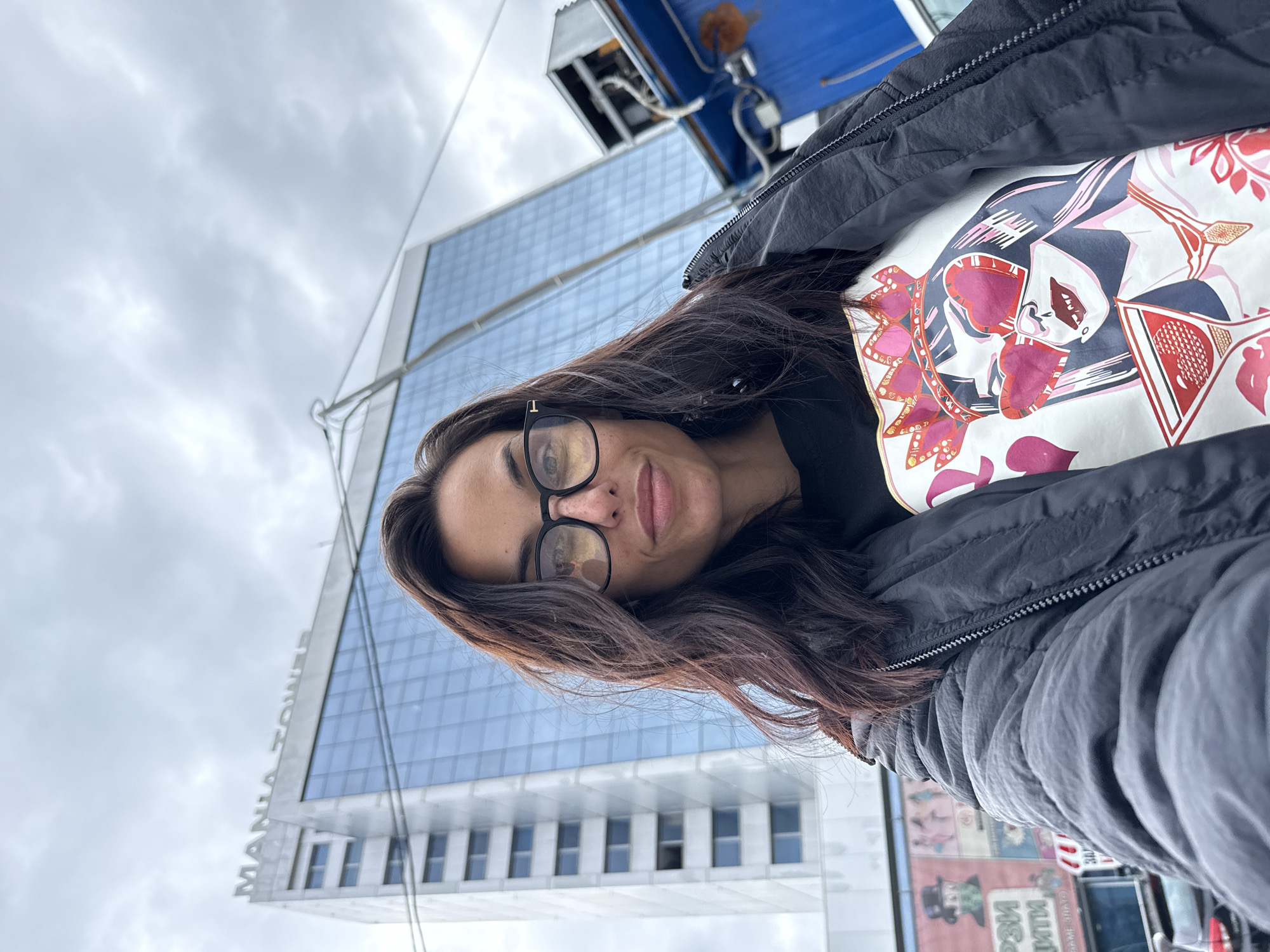 A woman with long, dark hair, wearing glasses and a black jacket, is taking a selfie in front of the Mania Tower in Pleven, Bulgaria. She is wearing a patterned shirt underneath her jacket. The sky is cloudy and the tower is visible in the background. There are other buildings and advertisements visible in the periphery of the image.