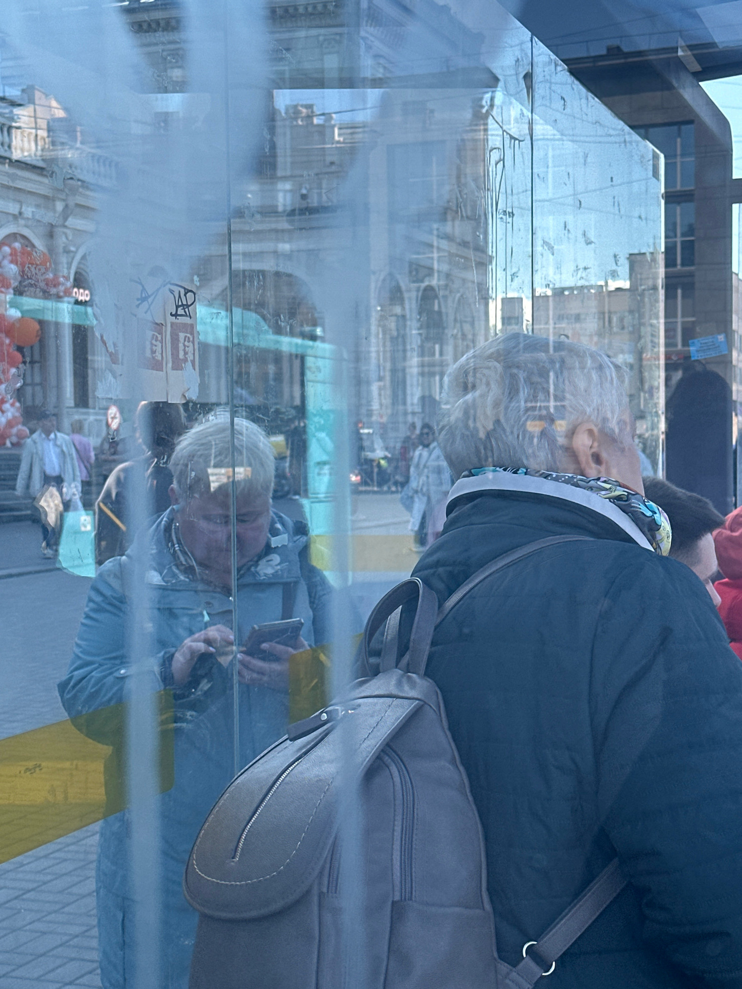 A close-up, slightly overexposed shot captures reflections on a glass surface, creating a layered and abstract depiction of a city scene in Saint Petersburg, Russia. In the foreground, the back of a person wearing a dark jacket and a grey backpack is visible. To the left, a woman in a blue jacket is reflected, looking down at her phone. Behind her, another reflection shows people and buildings. The overall impression is one of urban life viewed through a distorted lens, with architectural details of Saint Petersburg subtly present in the background. The lighting is bright, suggesting daylight, and the reflections create a sense of depth and complexity.