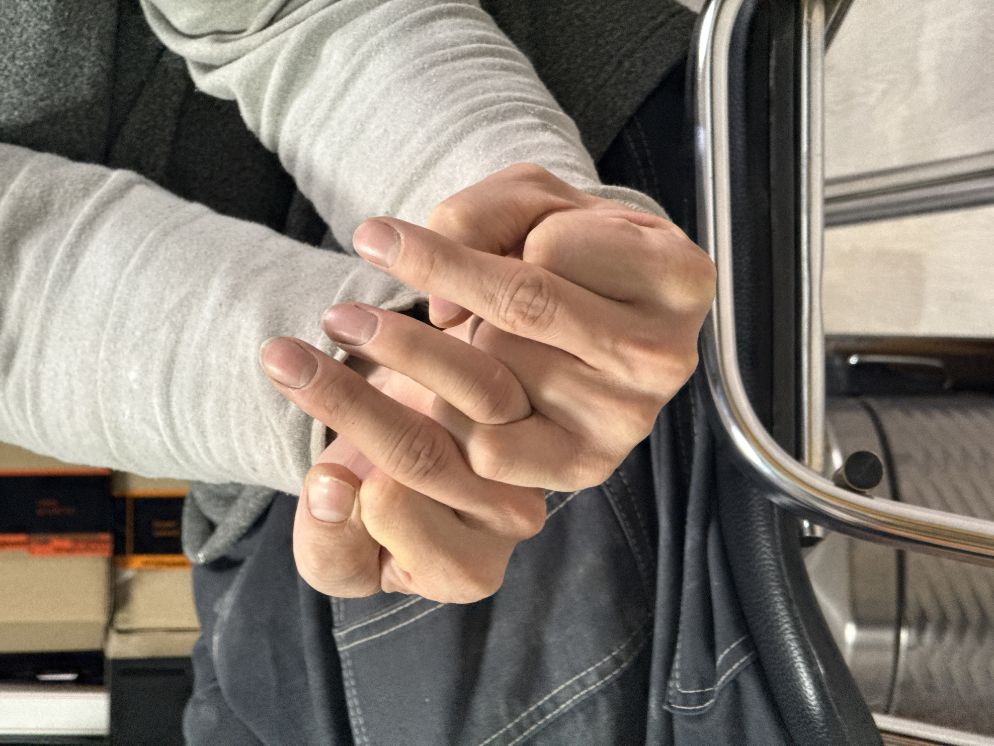 A close-up, eye-level shot captures a person's clasped hands in an indoor setting, likely in Chaikovsky, Russia. The individual, partially visible, wears casual clothing. Their forearms are covered by a light grey, subtly ribbed or textured long-sleeved garment, possibly a sweatshirt. Beneath this, a darker grey garment, perhaps a hoodie, is visible around the upper arm and shoulder area. On the person's lap or legs, dark grey or dark blue fabric with visible white stitching, characteristic of jeans or similar trousers, can be seen.
The person's hands are intertwined, with fingers gently interlocked. The fingernails are natural, without polish, and appear slightly uneven at the tips. The skin tone is fair, and some veins and knuckles are subtly visible, suggesting natural hand posture. The activity appears to be one of resting or contemplation, as the hands are held still.
To the left of the frame, the background is out of focus but shows a stack of what appear to be books or cardboard boxes, some with orange-brown details, implying an office, study, or storage area. To the right, a shiny, metallic, curved frame, likely part of a chair or furniture, is prominent. Behind this frame, a dark, possibly leather or upholstered surface is visible, along with a light-colored wall or surface further back.
The lighting suggests an indoor environment, possibly with ambient artificial light, as no strong shadows or directional natural light are evident. There is no visible text in the image.