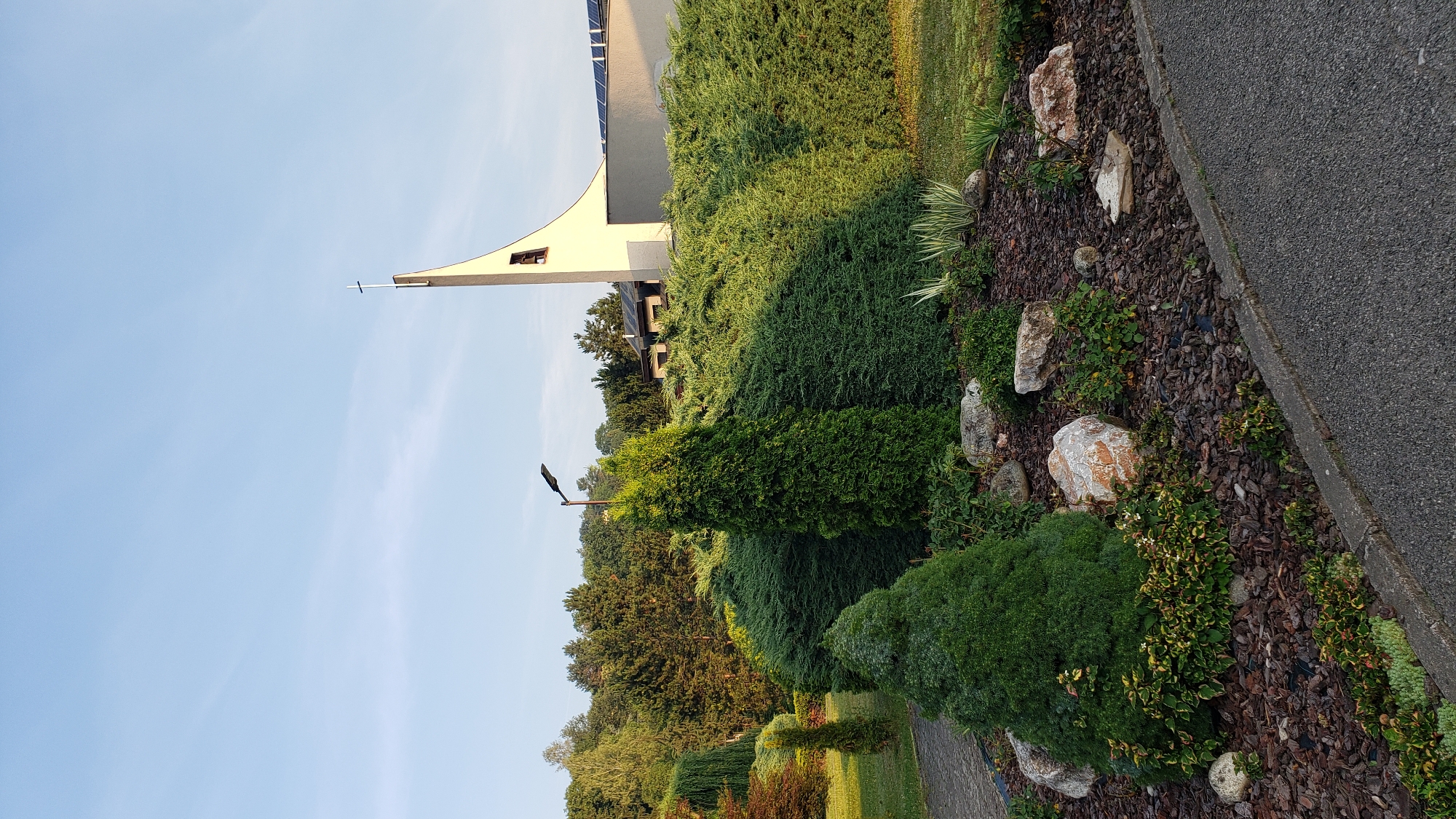 The image depicts the exterior of a modern church with a distinctive curved tower topped by a cross. The church is situated on a gentle slope, surrounded by well-maintained landscaping. In the foreground, there is a rock garden filled with mulch, various plants, and large, natural stones. Several evergreen trees and bushes, including a tall, conical cypress in the center and a rounded, dense shrubbery, are prominent. A lamppost stands behind the cypress tree. In the background, more trees and a building with solar panels on its roof are visible. The sky is clear and blue, indicating a sunny day. The overall scene is peaceful and suggests a well-kept church grounds. This church is located in the city of Prešov, Slovakia.