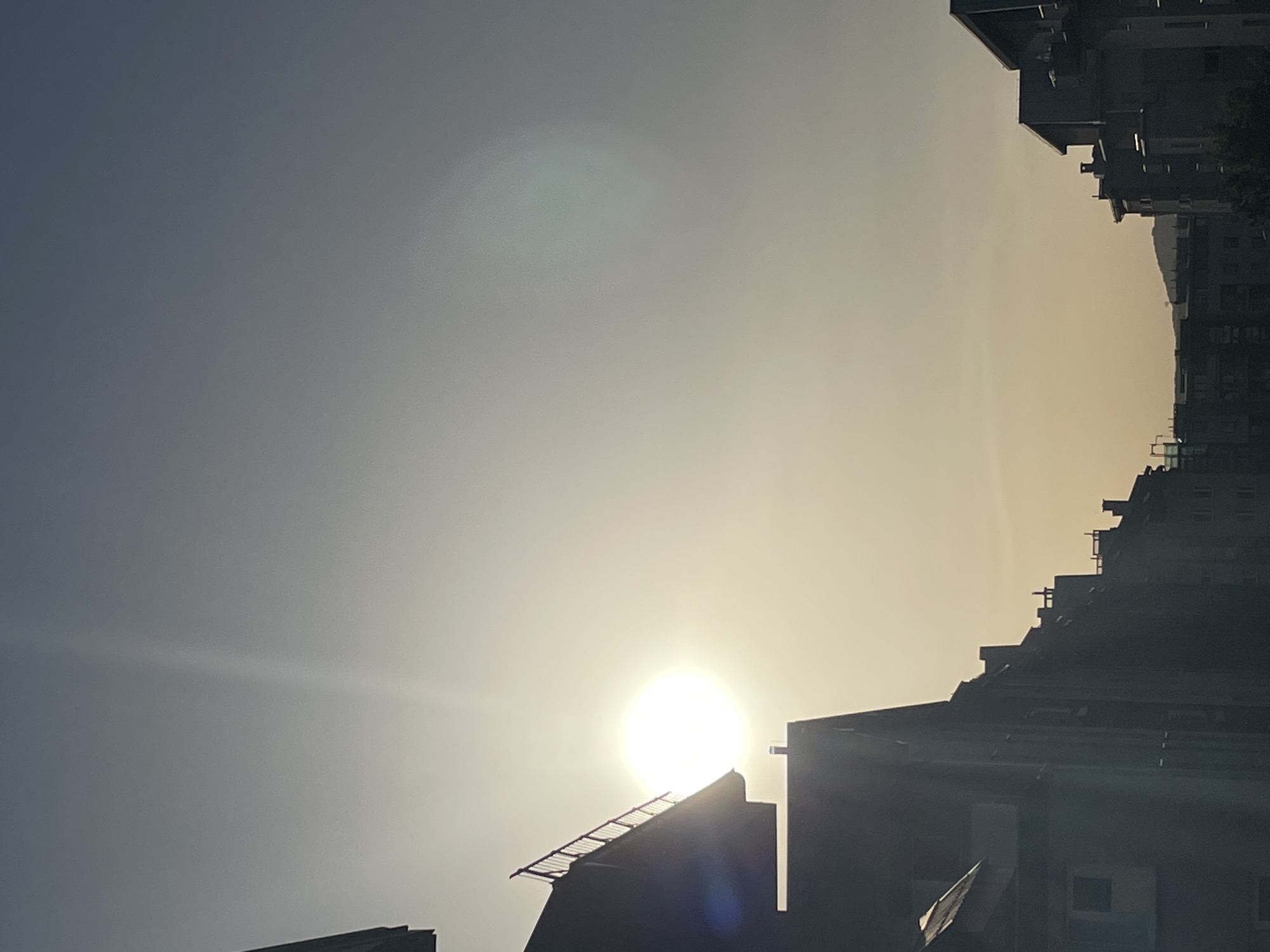 An extreme low angle shot shows the sun in a hazy sky shining between buildings. Silhouetted against the bright sun, the buildings are dark, and have a variety of architectural details including rooftops, balconies, and windows. The sky shifts in color from pale yellow near the sun to a gray blue in the upper left of the frame. The location is in the city of Unknown, Unknown.