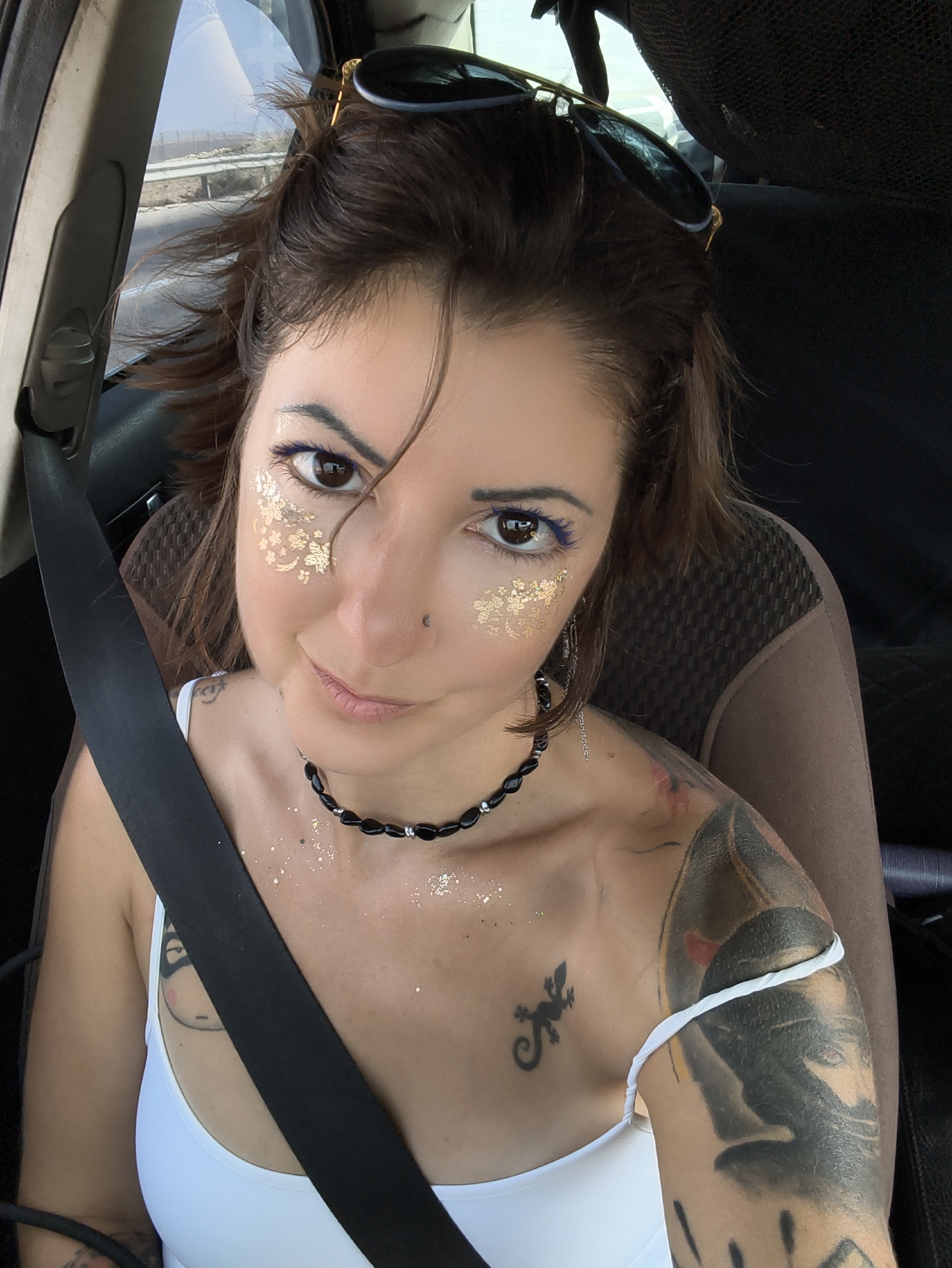 A medium shot captures a woman seated in a car's front passenger seat, facing the camera. She has brown hair and brown eyes, with purple eyeliner and gold glitter applied to her eyelids and cheeks. She is wearing a white tank top and a black beaded necklace. A black seatbelt is visible across her chest. Tattoos are visible on her left shoulder and arm. The background shows a view out of the car window of a road and landscape. The car appears to be in motion. The location is identified as the Ramat Negev Regional Council, Israel.