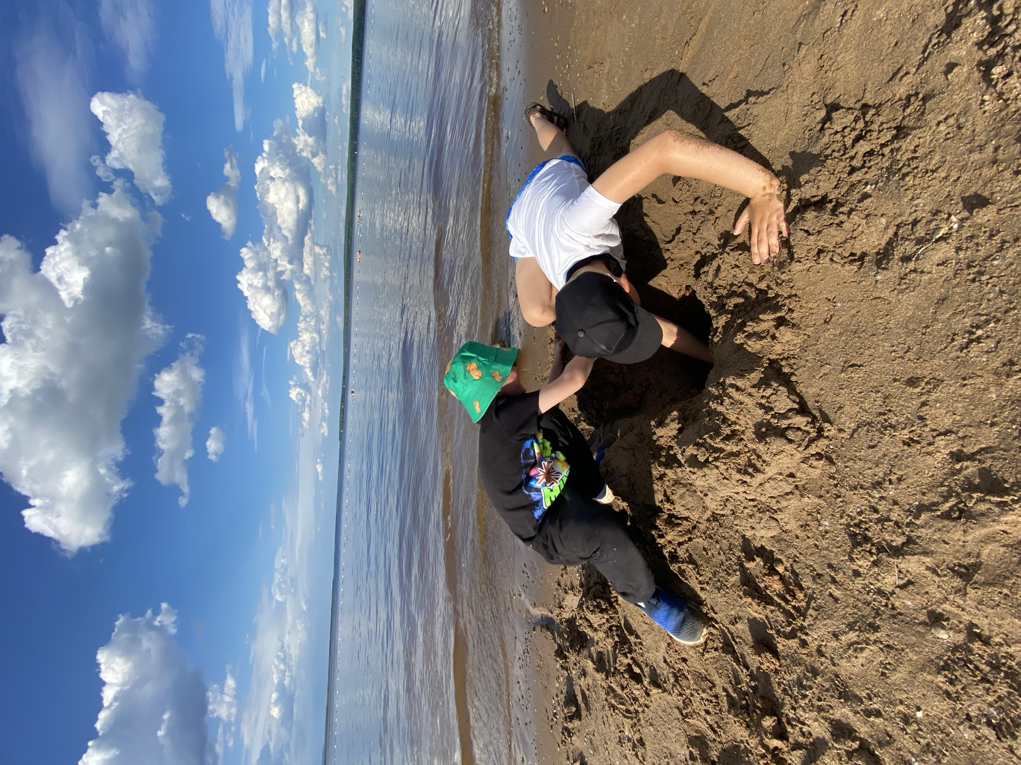 Two young males are digging a hole in the sand on the shore of the Kama River in Chaikovsky, Russia. The male on the left is wearing a black t-shirt, black pants, blue and black sneakers, and a green bucket hat. He is in a crouched position and appears to be using his hands to dig. The male on the right is wearing a white t-shirt, blue shorts, and a black baseball cap. He is lying on his stomach and reaching into the hole with his arms. The sand is wet near the water's edge, and the water is calm with a few boats visible in the distance. The sky is blue with scattered white clouds.