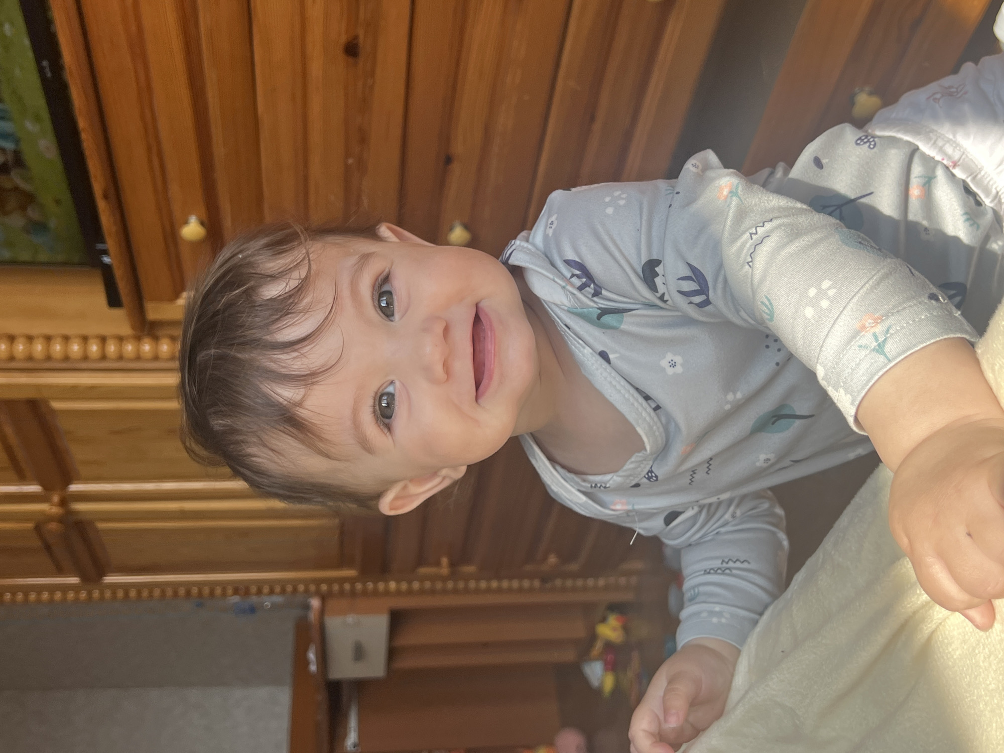 An infant, approximately 6-18 months old, with brown hair and dark eyes, is positioned in a low-angle, frontal close-up within a residential interior in the city of Elena, Bulgaria. The infant is smiling, with an open mouth revealing visible lower teeth, and looks directly towards the viewer. The infant is wearing a light-blue, long-sleeved onesie patterned with various small, multi-colored designs. The infant's left hand is gripping a cream-colored, textured blanket in the lower-left foreground.
The background consists of wooden furniture under strong natural light. To the infant's left, a tall wooden wardrobe with a vertical fluted trim is visible. Behind it, a light-colored wall, a small table, and a light-grey box are present. Several blurred, colorful objects, consistent with toys, are on the floor. To the infant's right, a wooden dresser with multiple drawers, including visible yellow and brown pulls, supports a flat-screen television displaying colorful animated content.