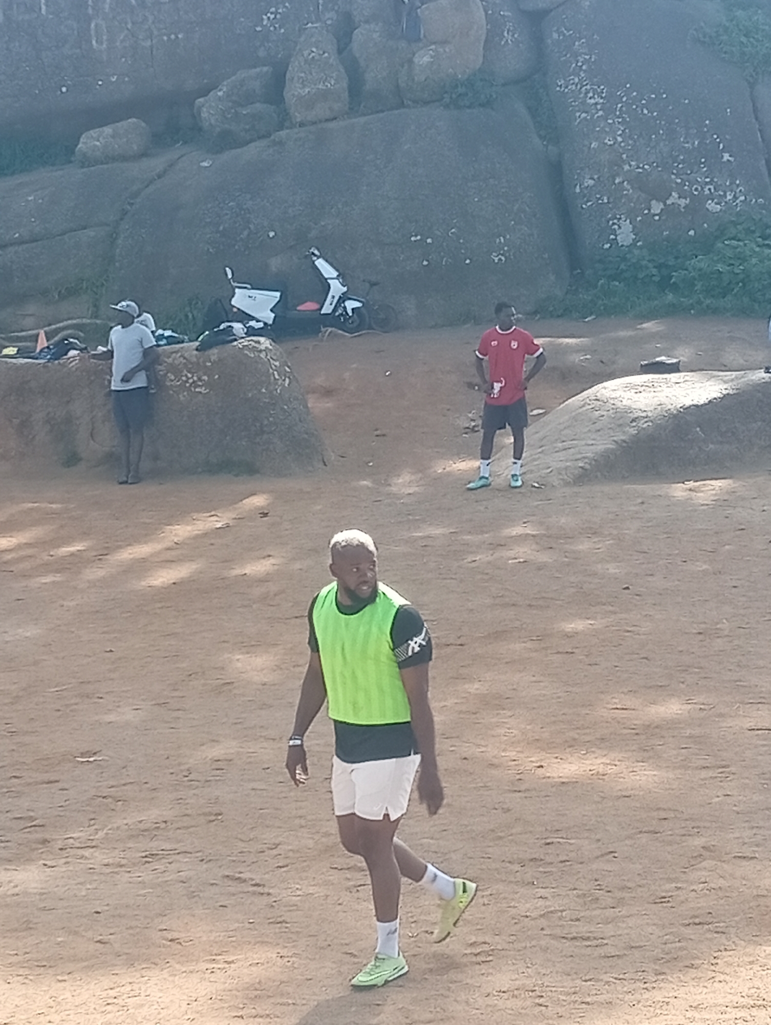The image depicts an outdoor scene in Jos, Nigeria, likely a training or recreational area. In the foreground, a man wearing a bright green vest over a dark t-shirt and white shorts walks on a dirt ground. He has short, grey hair and a beard. In the mid-ground, another man in a red t-shirt and black shorts stands with his hands on his hips, facing away from the camera. Behind him, to the left, a man in a grey t-shirt and dark shorts leans against a large rock formation. A white electric scooter is parked near the rocks. The background is dominated by a large, weathered rock outcrop, characteristic of the landscape in Jos. The ground appears to be sandy or dirt. The lighting suggests it is daytime, with shadows cast on the ground.