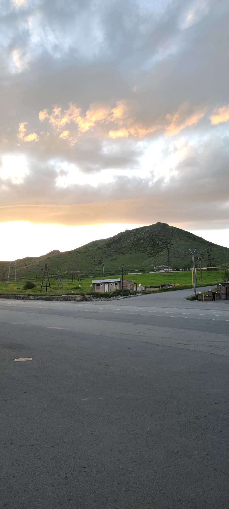 The image shows a landscape in Karadzor, Armenia, featuring a paved road in the foreground leading to a small building, a green hillside, and a mountain. Overhead power lines are visible traversing the hillside. The sky is filled with clouds illuminated by a setting or rising sun. A person is visible on the right side of the image, standing next to the road. The road surface is gray asphalt.