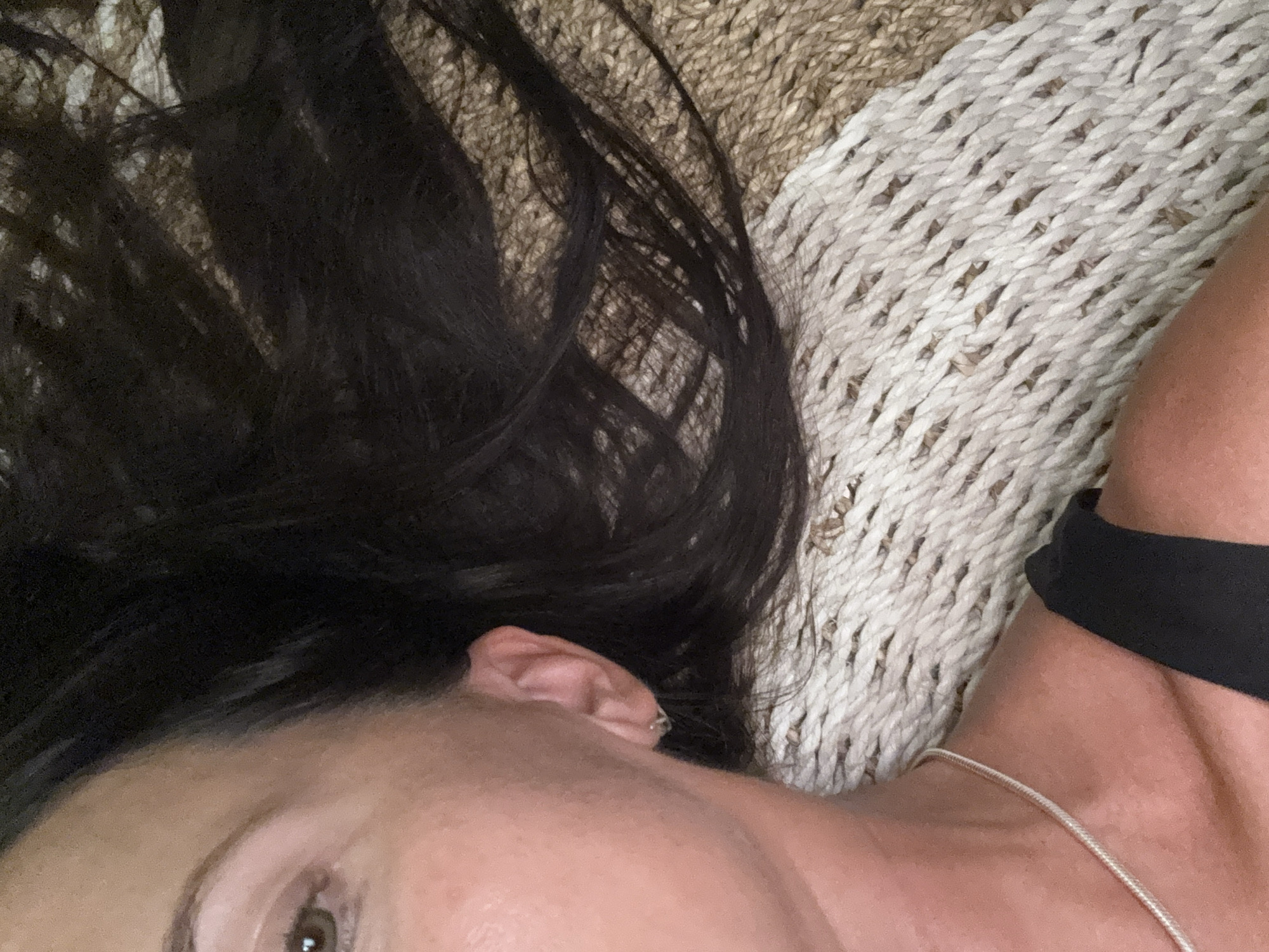 A close-up, diagonally oriented image captures a person lying down, predominantly showing the left side of their upper body. The subject's dark, long hair is spread across a textured background, which appears to be a woven mat or rug. This mat features two distinct sections: one in natural brown and tan tones, and another in off-white or cream, both exhibiting a braided or knotted pattern.
Visible features of the person include a portion of a brown eye, an ear adorned with a small silver hoop earring, and the cheek and jawline. A delicate silver chain necklace rests on their neck, which shows warm skin tones. Part of a black strap, likely from clothing, is visible over the right shoulder. The person appears to be in a state of rest or relaxation. The lighting is soft and even. No specific activities are depicted beyond this resting posture. There is no visible text within the image. This scene is set in Kuta Selatan, Indonesia.