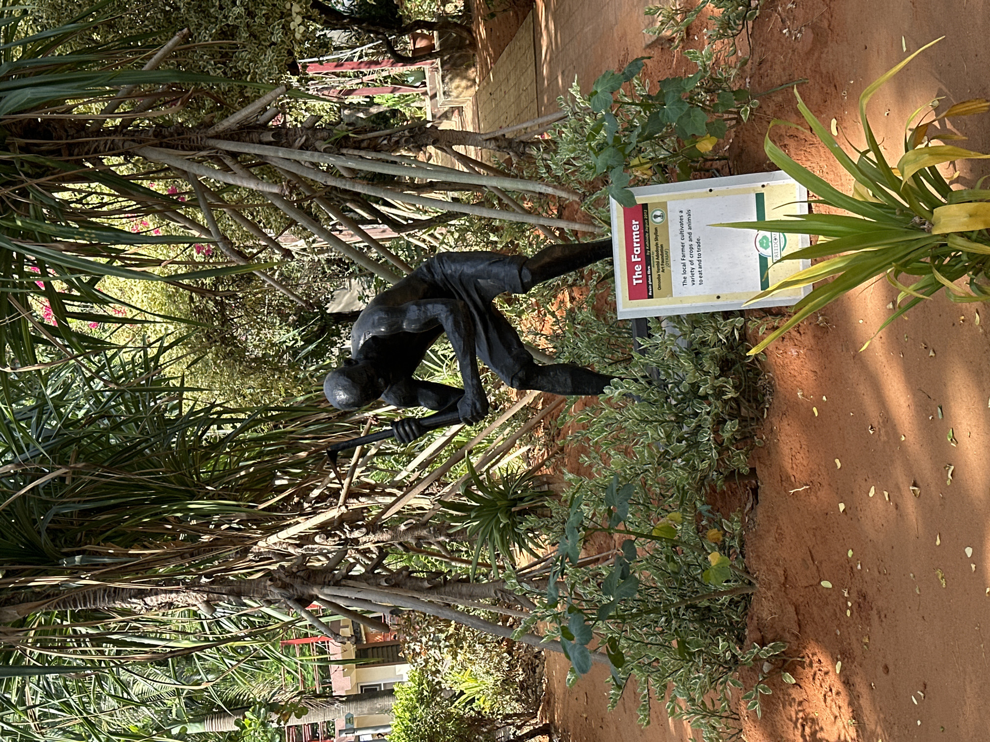 The image shows a bronze sculpture of a farmer tilling the soil, surrounded by greenery in a park or garden setting. A sign next to the sculpture identifies it as "The Farmer" by Kehinde Jagun (2012), a resin glass fibre work from the Omooba Yemisi Adedoyin Shyllon Art Foundation (OYASAF). The sign explains that the local farmer cultivates a variety of crops and animals to eat and to trade. The ground is reddish-brown, indicating the soil type. The sculpture and sign are partially obscured by trees and bushes. Given the mention of the OYASAF, a foundation known for promoting Nigerian art, and the artist's name, the location is likely Lagos, Nigeria.