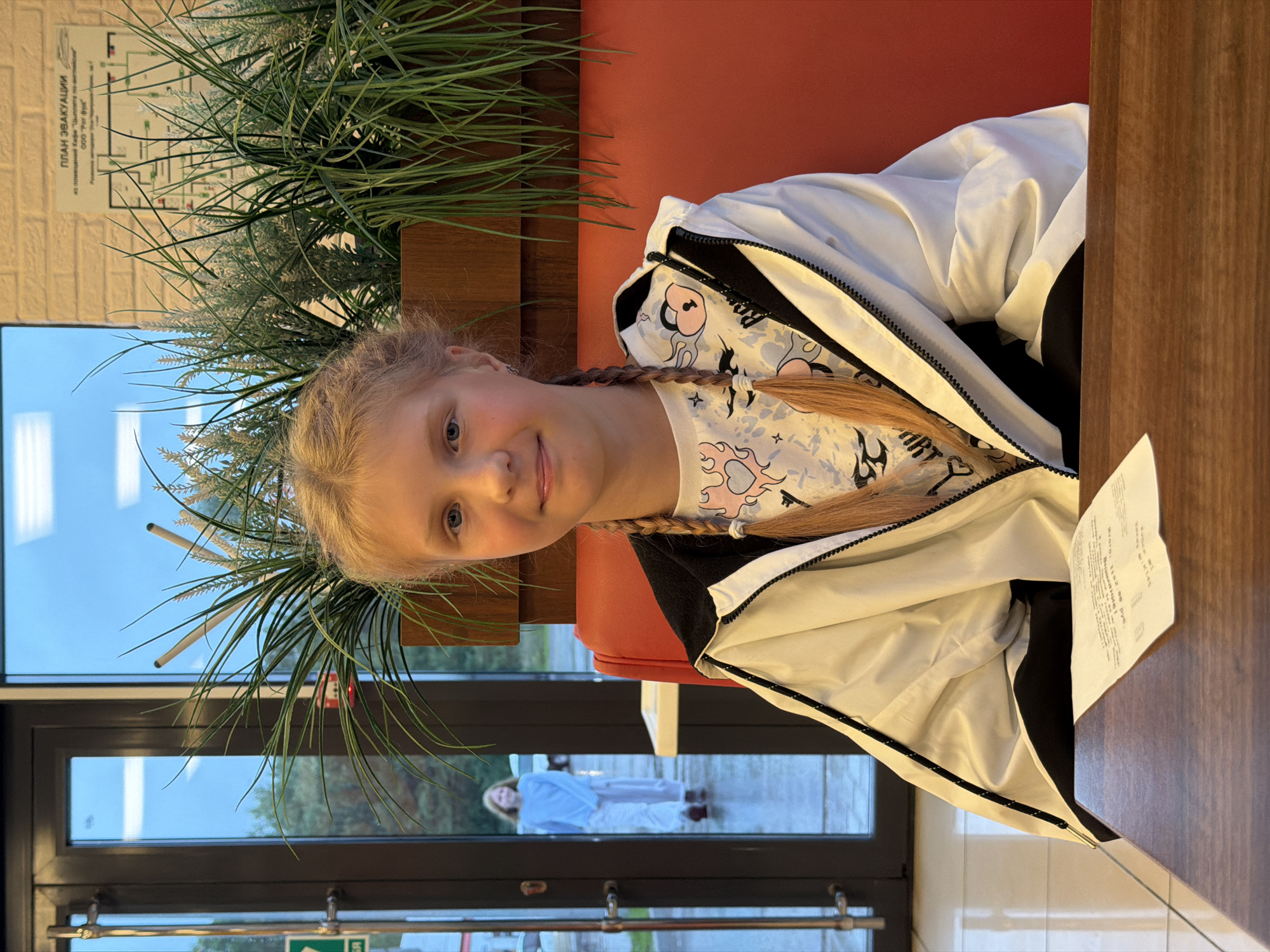 A young girl with blonde hair in two braids is sitting at a table, facing the camera. She is wearing a white jacket with a black zipper and a t-shirt with a graphic print. Behind her, there is a red seat and a window with a view of what appears to be a natural landscape or park. To the left of the girl, there is a large potted plant with long, spiky leaves. A paper with some text is visible on the right side of the image. The overall scene appears to be indoors, possibly in a cafe or restaurant, given the table and seating. The lighting suggests it could be daytime, with the sky visible through the window indicating an outdoor setting. The girl's expression is neutral.
A sign with Cyrillic text is visible on the wall behind the plant. The text, in part, reads "ПЛАН ЭВАКУАЦИИ" which translates to "EVACUATION PLAN".
Through the window, another person is visible outside, wearing a light-colored outfit and standing on a paved area. The trees and greenery suggest an outdoor location.