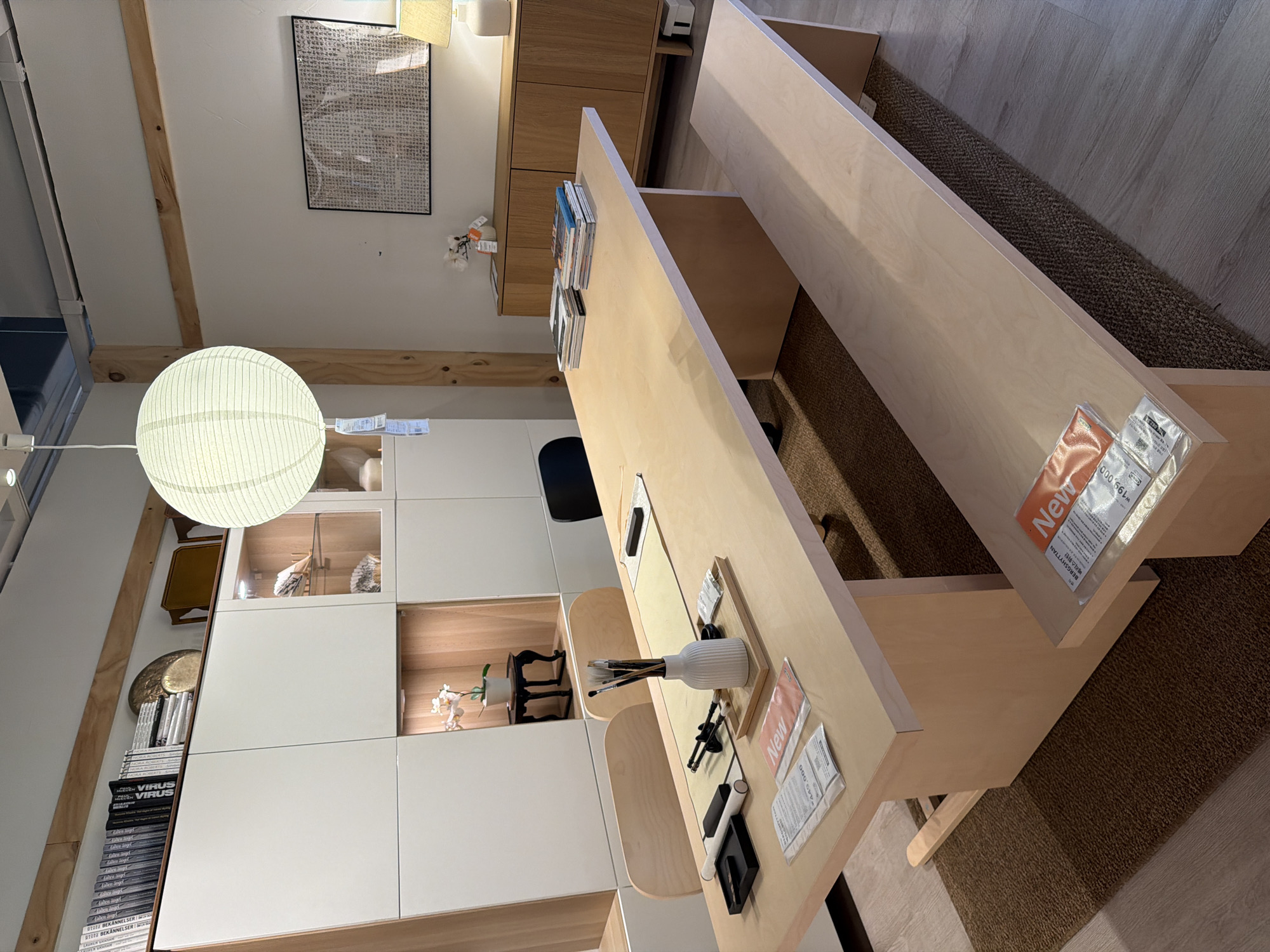 The image depicts a bright, modern room, likely a showroom or a well-lit living space. A long, light-wood dining table with a matching bench is the central focus. On the table are art supplies, including brushes in a white vase, and several magazines. Behind the table, a row of white modular cabinets lines the wall, some with glass fronts showcasing decorative items and a potted orchid. To the right of the cabinets is a wooden sideboard with a lamp and a framed piece of artwork above it. A large, circular white paper lantern hangs from the ceiling, illuminating the space. The flooring is a light wood laminate, with a textured brown rug under the table and bench. The scene is clean and uncluttered. The location is identified as Goyang-si, South Korea.