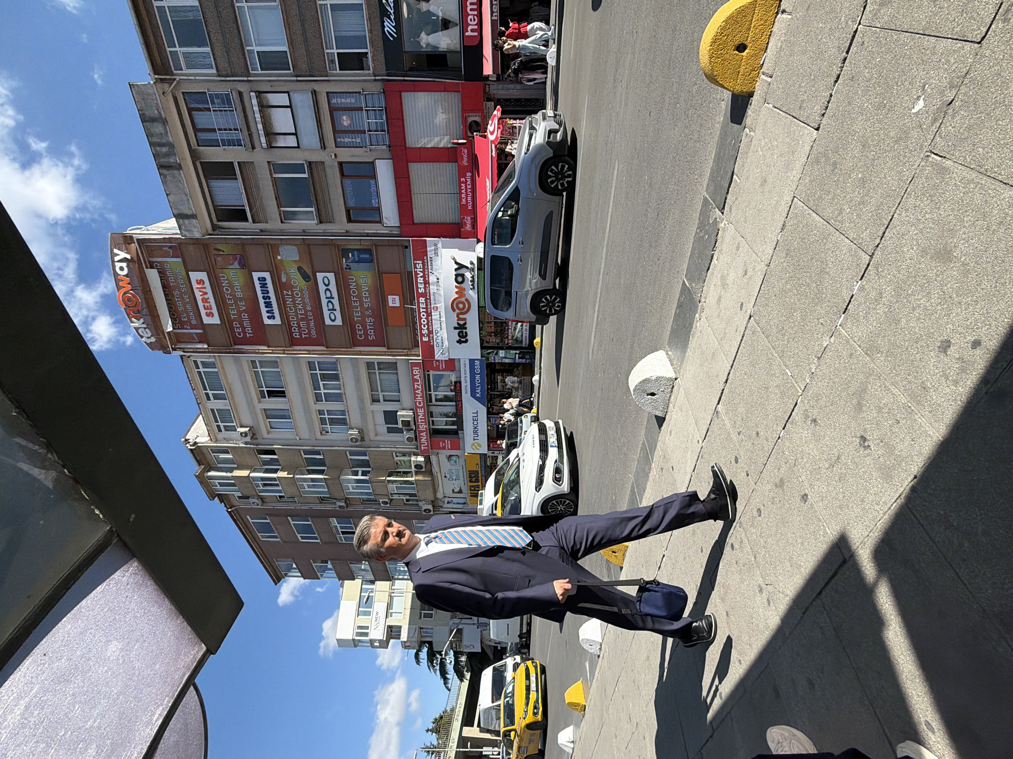 In Kadıköy, Turkey, a man in a dark navy suit, white shirt, and a blue and white striped tie walks on a sidewalk. He is carrying a dark blue shoulder bag in his right hand. He is walking towards the right of the frame. The street has a yellow taxi and a silver van driving on it. Buildings line the street, with various signs visible, including "tekroway," "SAMSUNG," "OPPO," and "TUNA İŞTİHMAZ CLAZERİ." The sky is blue with some clouds. The sidewalk is made of square concrete tiles, and there are several concrete and yellow bollards along the edge of the street. The scene appears to be a daytime urban environment.