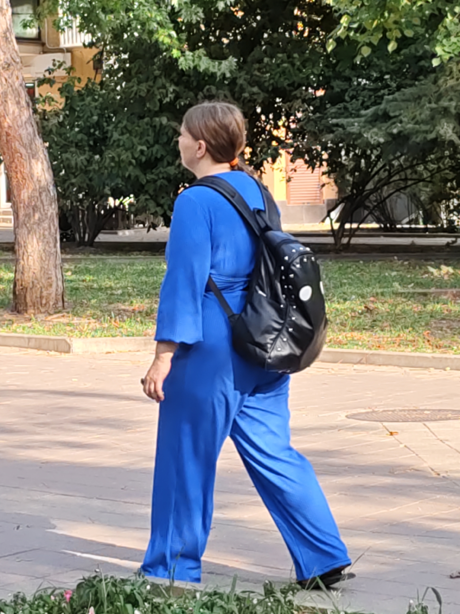 A middle-aged woman with brown hair pulled back is walking on a paved path in a park-like setting. She is wearing a bright blue, wide-legged jumpsuit with bell sleeves and carrying a black backpack decorated with studs and white circular patterns. She is looking to her left. Behind her, a tree trunk is visible on the left, and lush green trees and bushes line the background. A building with beige siding and windows is also partially visible behind the foliage. The ground is paved with light-colored rectangular stones. The scene appears to be in Rostov-on-Don, Russia.