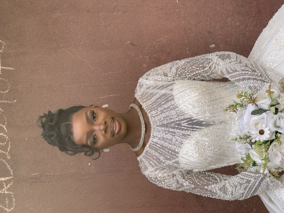 A young adult Black woman stands facing the camera, positioned centrally against a textured, reddish-brown wall. Her dark hair is styled in an elaborate updo with curls framing her face. She wears full makeup, including defined eyebrows and a subtle smile. She is adorned with a double-strand choker necklace and rectangular drop earrings, both featuring small, clear stones. She is wearing a long-sleeved white wedding gown, intricately embellished with clear beads and sequins that form patterns across the bodice and sleeves. The gown features an illusion neckline over a sweetheart cut. In her arms, she holds a bridal bouquet composed of white roses, white daisies, and various green foliage. Faint, faded white markings, possibly chalk or paint, are visible at the top edge of the wall behind her, appearing to spell "EAD 2020 TO". The scene is located in Garaku, Nigeria.