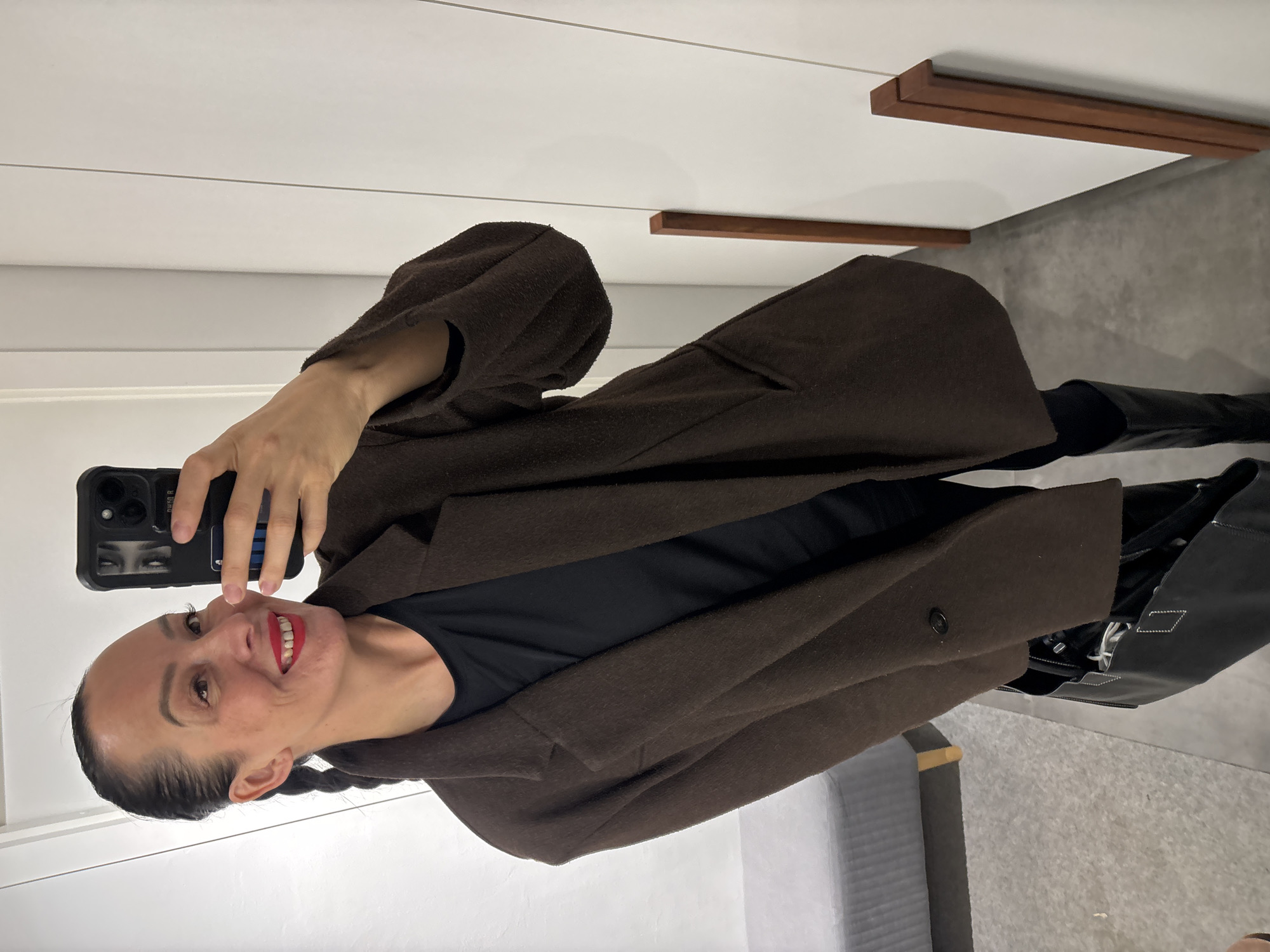 A woman with dark hair in pigtails, wearing a brown coat over a black top and tall black boots, takes a selfie in what appears to be a dressing room. The phone case has a design of eyes on it. The background includes light-colored walls with horizontal lines and wooden fixtures, as well as a gray floor. There is a black bag in the lower right corner. It's not possible to determine the location in Chaikovsky, Russia from this image alone.