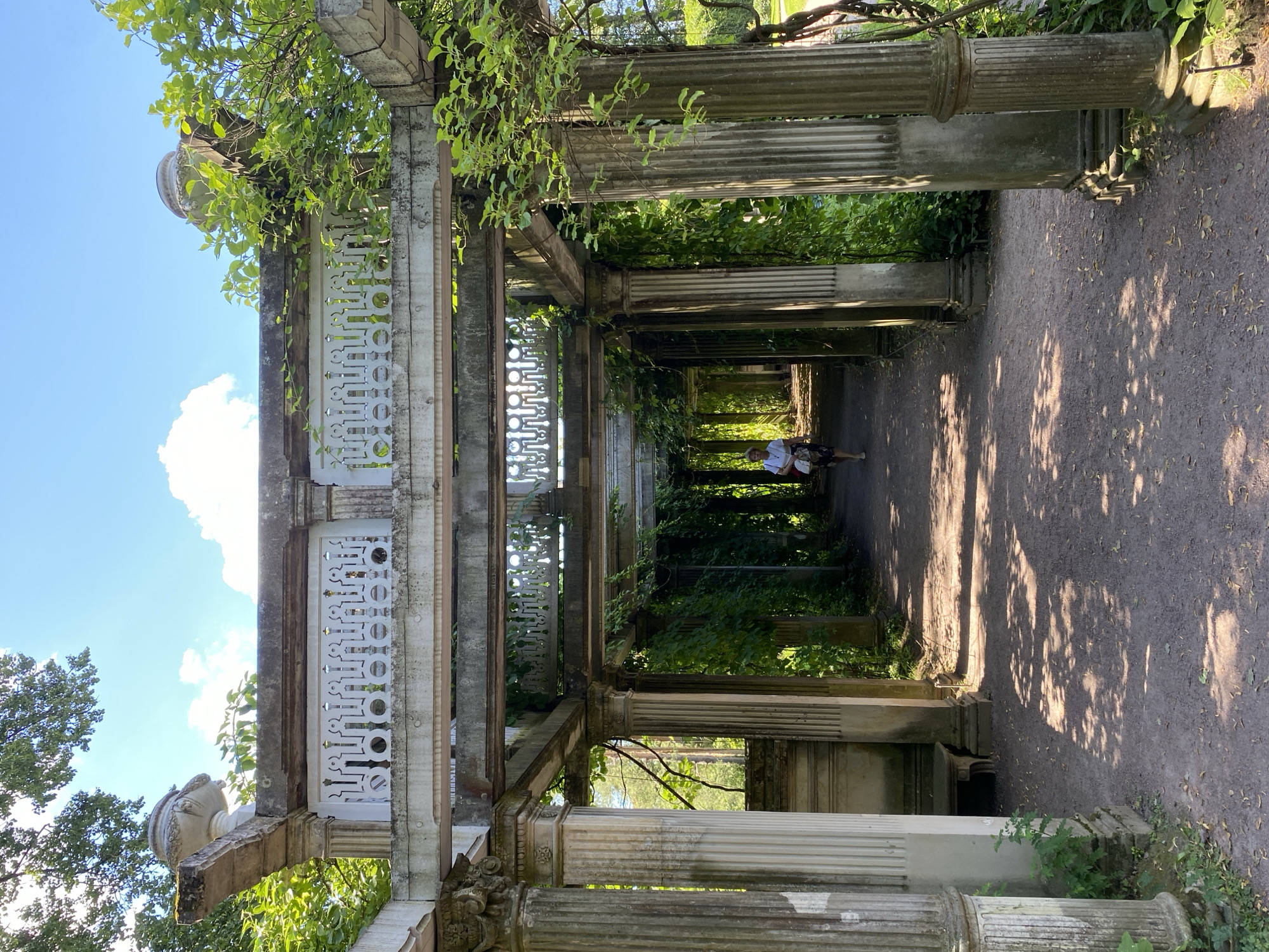 The image depicts a woman walking down a pathway through a stone pergola in the city of Pushkin, Russia. The pergola is supported by fluted columns and features ornate white railings. Lush green ivy and other foliage are growing on and around the structure, creating a shaded, natural atmosphere. Sunlight dapples the gravel path, casting shadows from the overhead branches. The woman, wearing a white top and patterned skirt, is seen from the side, walking away from the camera into the dappled sunlight. In the background, trees and a bright blue sky with scattered clouds are visible. The overall impression is one of a serene, historical garden setting.