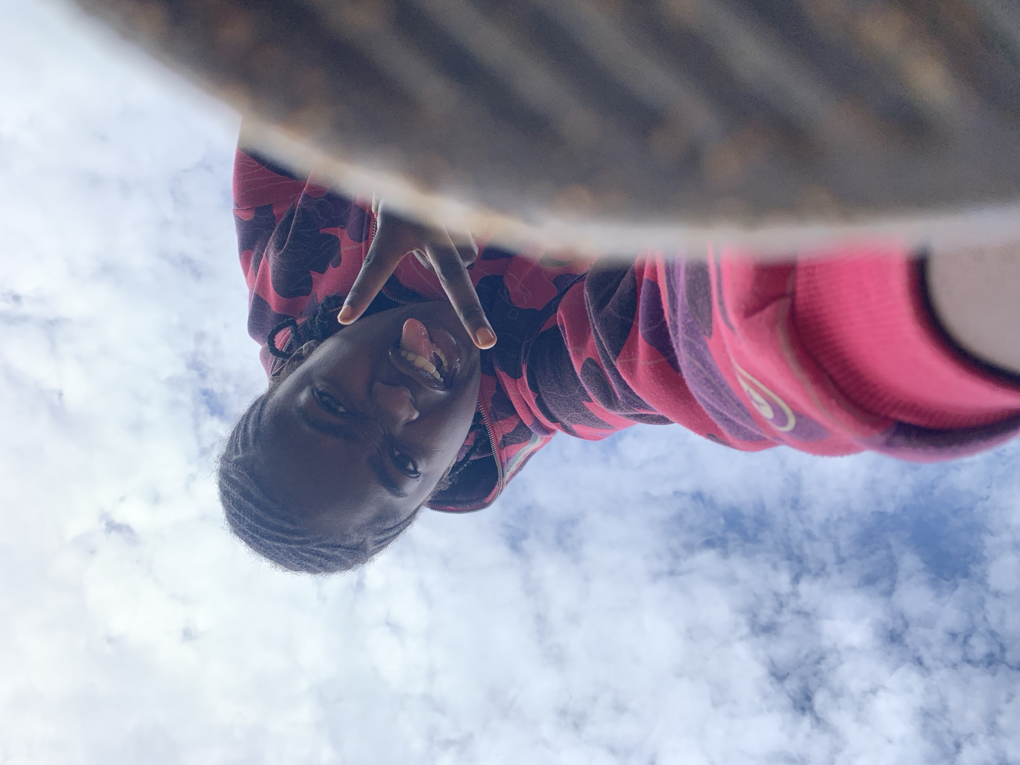 This image, purportedly taken in Rafin Par, Nigeria, displays an individual from a low-angle perspective, looking downwards towards the camera against a cloudy sky. The subject, exhibiting a dark complexion and hair styled in multiple tight braids, is wearing a hooded sweatshirt featuring a red and dark-patterned camouflage design on the upper body and bright pink on the lower sections and sleeves. A light-colored, curved emblem, resembling a logo, is partially visible on the pink sleeve. The individual's facial expression includes an open mouth with the tongue slightly extended, and a peace sign (V-sign) is formed with the fingers of their left hand, positioned near their face. The background consists primarily of white and grey clouds with discernible patches of blue sky. A large, out-of-focus object, characterized by a mottled brown texture, occupies the right side of the frame, obscuring a significant portion of the subject's body.