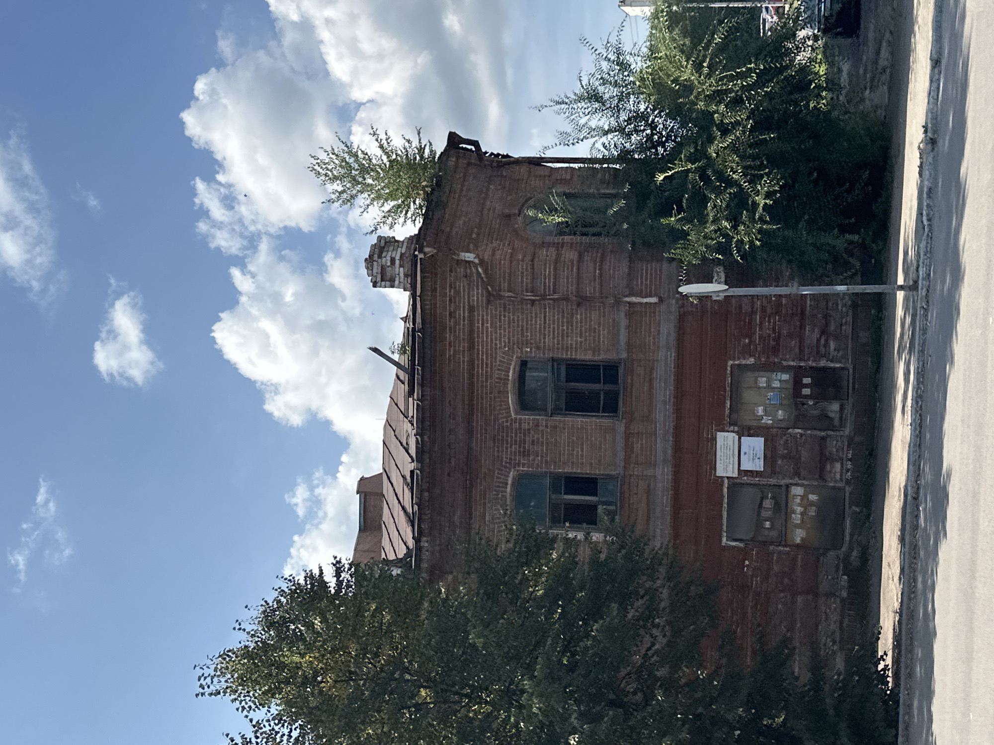 The image depicts a dilapidated brick building, likely an old residential or commercial structure, located in Biysk, Russia. The building features red brick construction, arched windows, and a damaged, sloping roof with visible signs of disrepair, including missing shingles and what appears to be a tree growing from the roofline and another from the chimney. The ground level of the building has several doors and windows, some boarded up or covered with notices. Overgrown greenery, including trees and bushes, surrounds the building, partially obscuring its facade. The scene is set against a backdrop of a blue sky with scattered white clouds, suggesting daytime. A paved road and sidewalk are visible in the foreground. A metal signpost stands on the sidewalk near the building. In the background, the roofline of another building and parts of parked vehicles can be discerned. The overall impression is one of neglect and age.