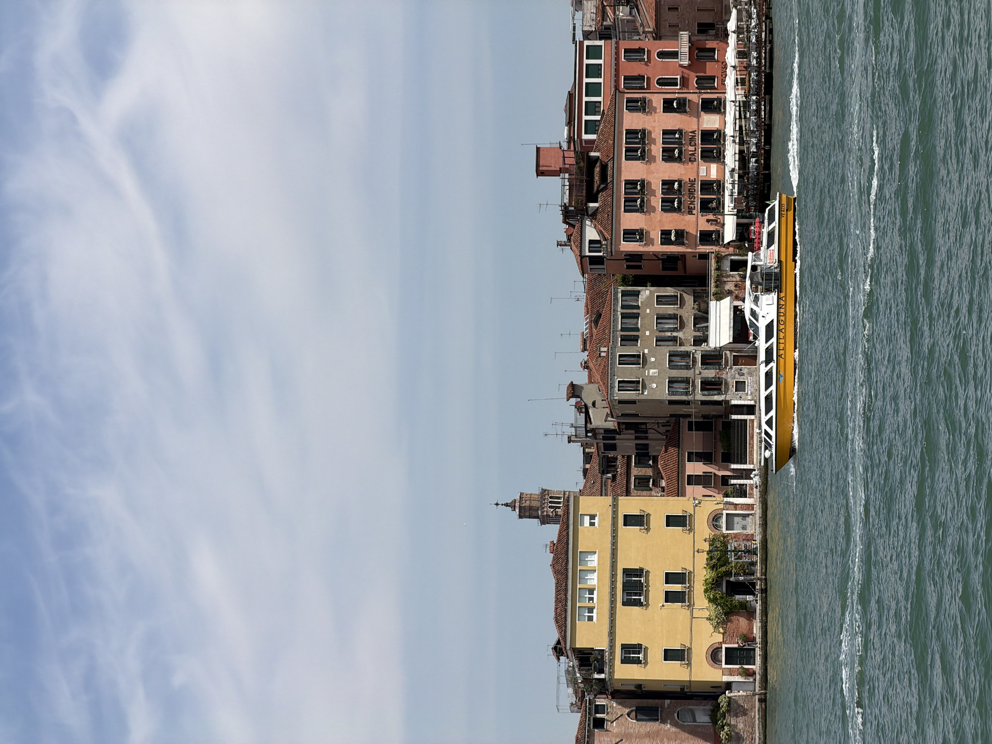 A vertical, eye-level photograph captures Venice, Italy, along a waterway. The buildings in various architectural styles are adjacent to the water. A yellow and white water bus is docked or slowly moving along the waterway's edge. The sky is pale blue with soft, wispy clouds. The water is a mottled grey-green, indicating movement or depth. The buildings are several stories high and vary in color, including yellow, brown, and red. The image provides a clear, realistic depiction of urban architecture and marine activity in Venice.