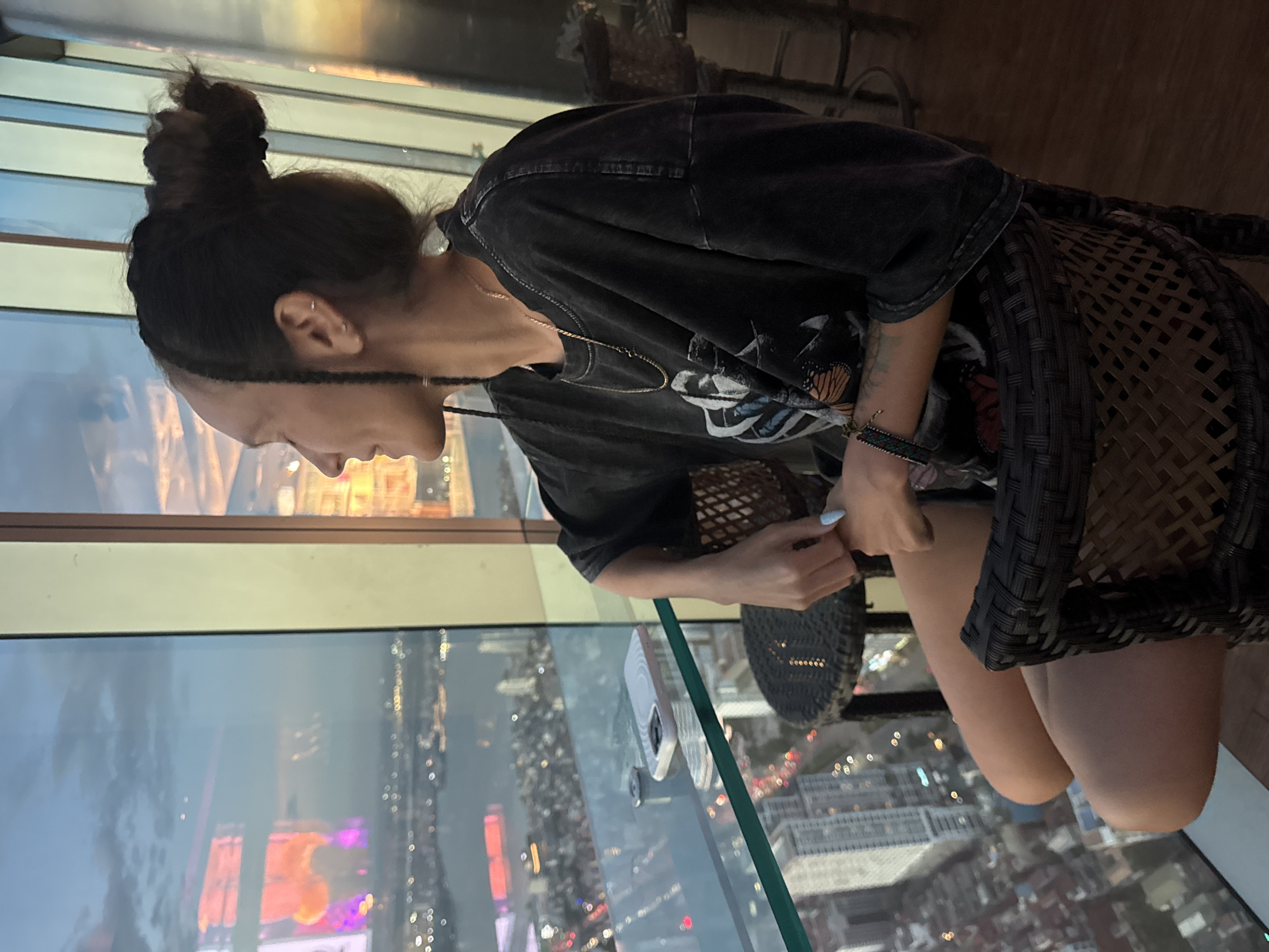 A person sits in a chair at a table by a large window, overlooking a city at dusk. The person, with dark hair in a bun and braids, wears a dark t-shirt with a graphic print and a necklace. They are looking out the window at the city lights and the sky. On the glass table in front of them is a phone. The city appears to be Hanoi, Vietnam, based on the distinctive architectural style visible in the background and the presence of the Lotte Center Hanoi, a prominent skyscraper in the city. The perspective from a high vantage point suggests the location is likely a bar or restaurant on an upper floor of a building.
