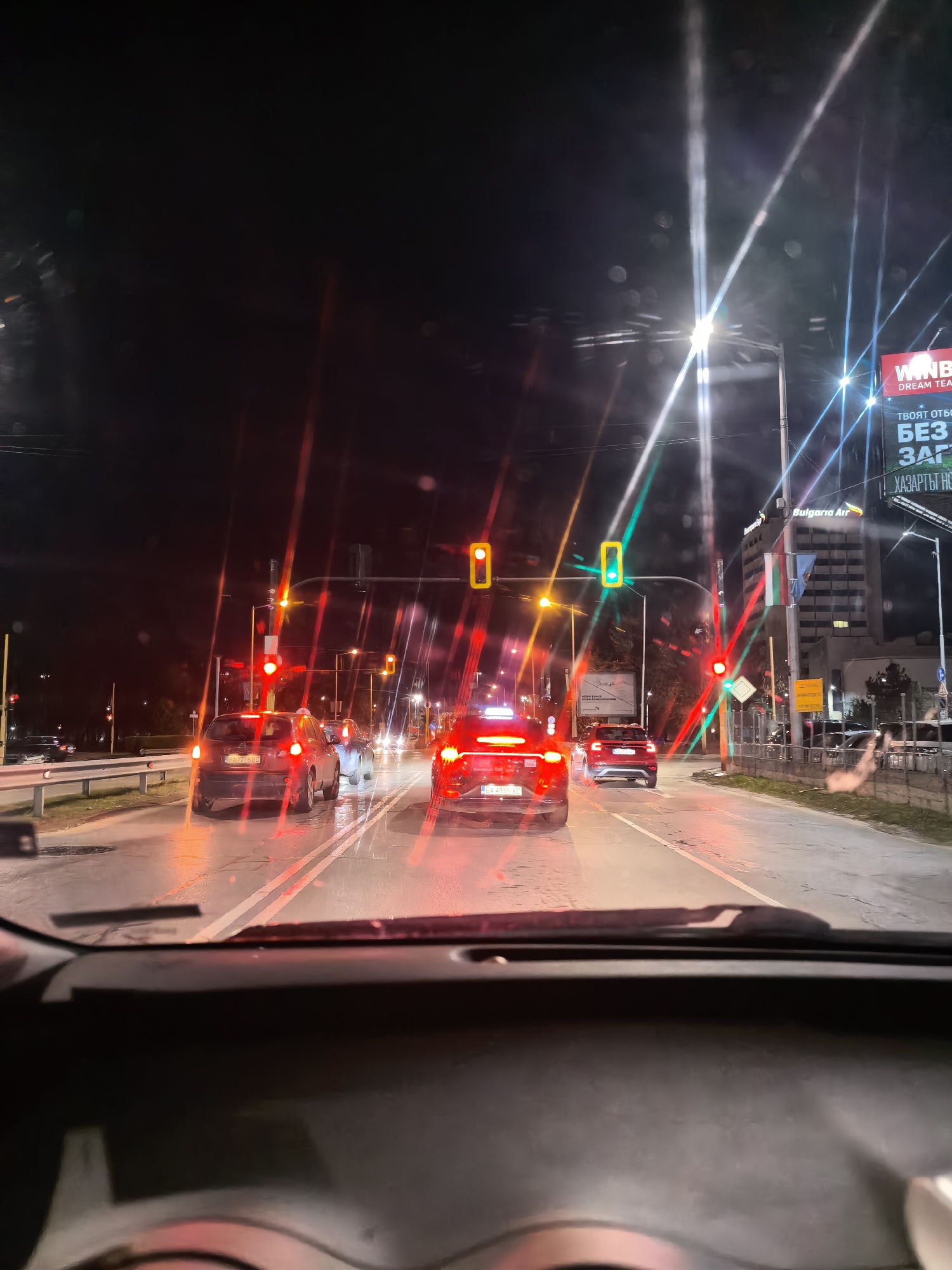 Here is a detailed description of the image:
The image is taken from inside a car at night, showing a view of a road with several cars and traffic lights. The car's dashboard and steering wheel are visible at the bottom of the frame. The traffic lights are red, with one showing green, indicating a stop condition for the car taking the photo. The road appears wet, reflecting the lights from the cars and street lamps. The cars are positioned in lanes, waiting at the traffic lights. In the background, there are buildings and a large billboard with the word "WINB" and other text in a foreign language, along with the name "Bulgaria Air". Flags of what appears to be Bulgaria can be seen near a tall building. The overall lighting conditions suggest it is nighttime in a city environment.