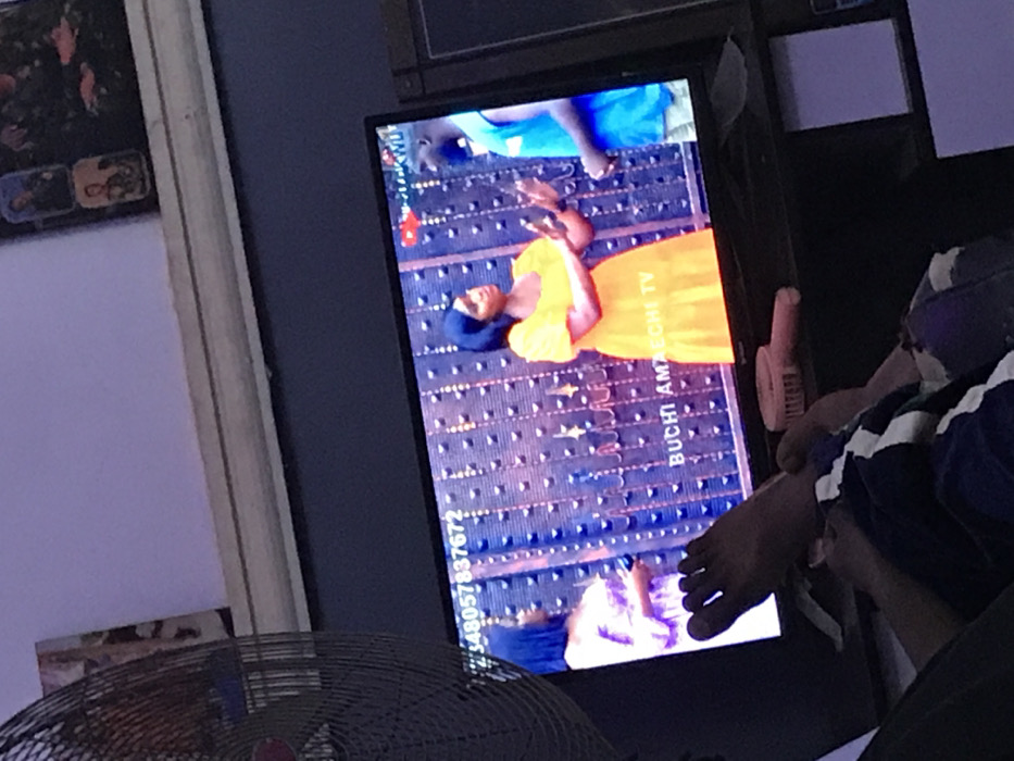 A television displays a program featuring two individuals on a patterned carpet. One person wears a yellow dress and the other is in blue. The screen displays "BUCHI AMAECHI TV" and a phone number. A foot rests near the screen. In the foreground, a fan is partially visible. The overall image appears casually taken indoors.