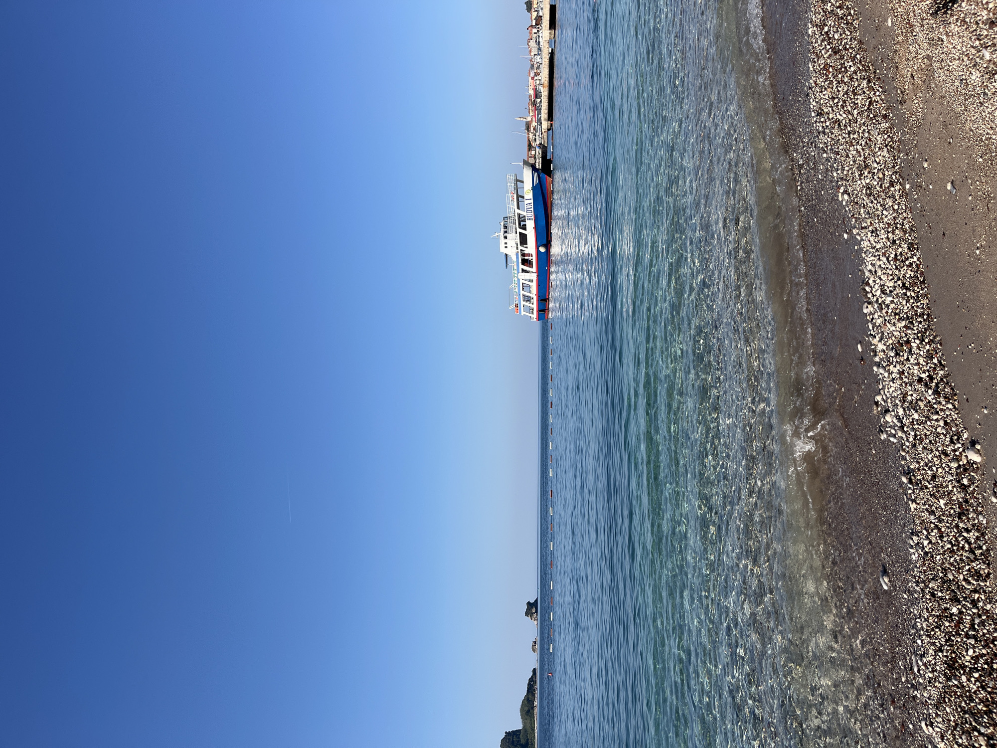 The image captures a serene coastal scene in Budva, Montenegro, under a clear, bright blue sky. The foreground features a pebbly beach with small waves gently lapping at the shore, revealing the transparent, shallow water. In the middle ground, the calm sea stretches towards the horizon, marked by a line of buoys. A white and blue passenger boat, identified by the lettering "BUDVA I" on its side, is docked at a pier in the middle right of the frame. Beyond the boat, a town with red-tiled roofs and a church spire is visible, suggesting the urban landscape of Budva. On the left side of the image, a rocky island formation rises from the sea, with lush greenery on a distant headland. A few people are visible near the pier, and a single contrail streaks across the expansive sky.