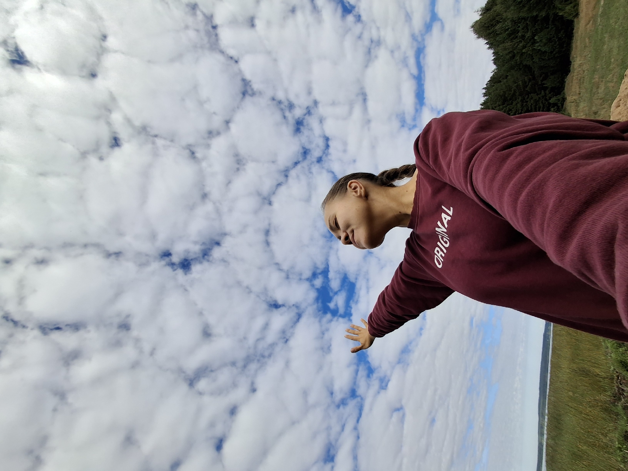 A young woman with her eyes closed and her arms outstretched stands in a field, looking up at a cloudy sky. She is wearing a maroon sweatshirt with the word "ORIGINAL" printed on it in white letters. The background shows a body of water and a line of trees. The image appears to be taken in Plishkino, Russia.