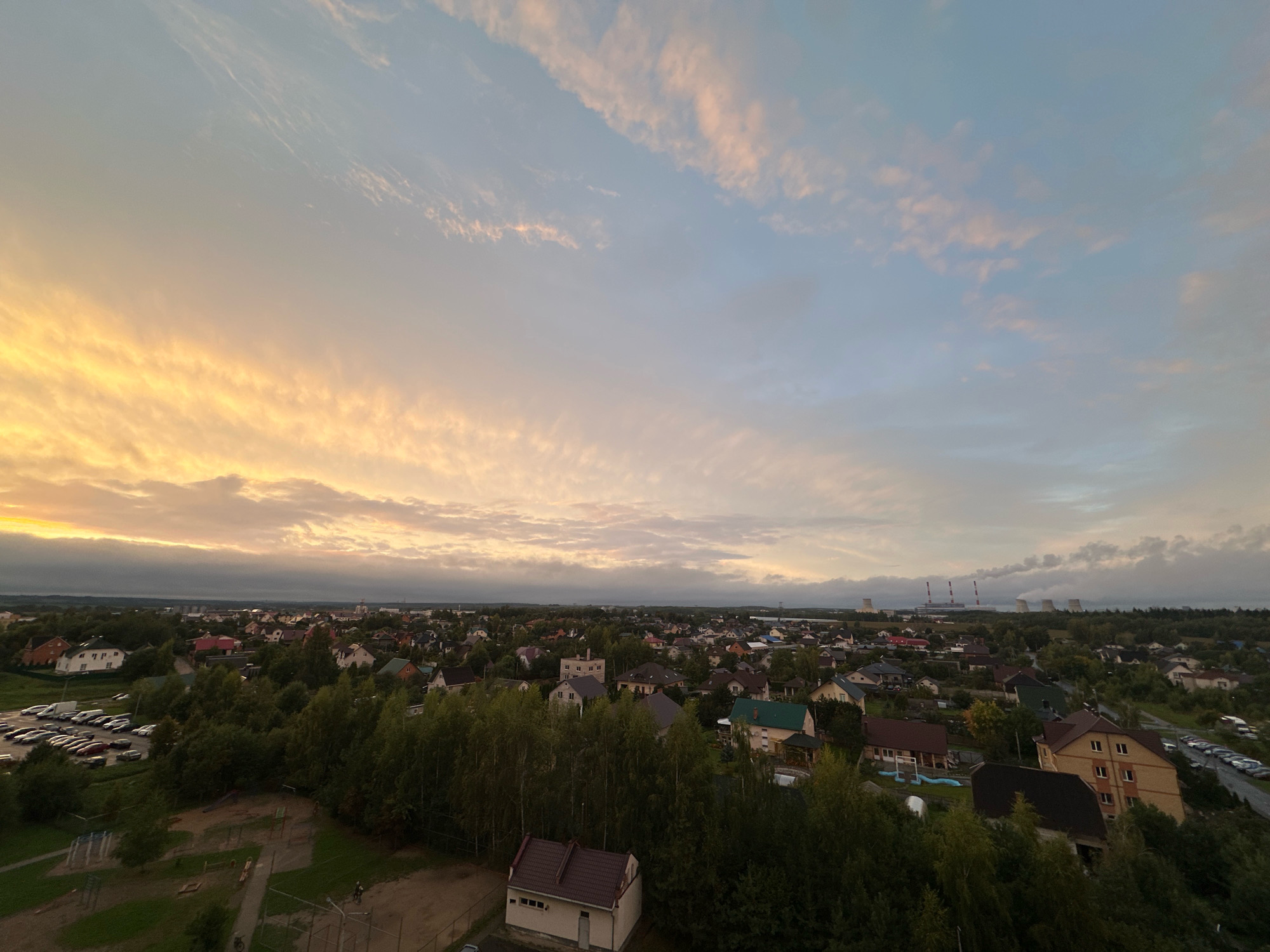 This is an aerial, wide-angle photograph of a residential area on the outskirts of the city of Shchomyslitsky Selsoviet, Belarus, during a sunset. The sky dominates the upper portion of the frame, filled with a gradient of colors from warm golden yellows and oranges on the left, transitioning to soft pinks and blues on the right, with wispy clouds scattered throughout. Below the sky, a suburban landscape unfolds. Dense green trees and foliage form a prominent element, interspersed with numerous houses of varying architectural styles and colors. In the mid-ground and background, more houses are visible, some with red or brown roofs. Further in the distance, industrial structures with tall, smokestack-like elements emitting plumes of vapor are visible on the horizon, suggesting a power plant or industrial complex. In the foreground, elements of a park or recreational area are present, including a playground with swings and sandboxes, and pathways. A parking lot filled with several cars is visible on the left side of the frame. The overall scene depicts a tranquil evening in a suburban setting, with the natural beauty of the sunset contrasting with the presence of human habitation and industrial activity.