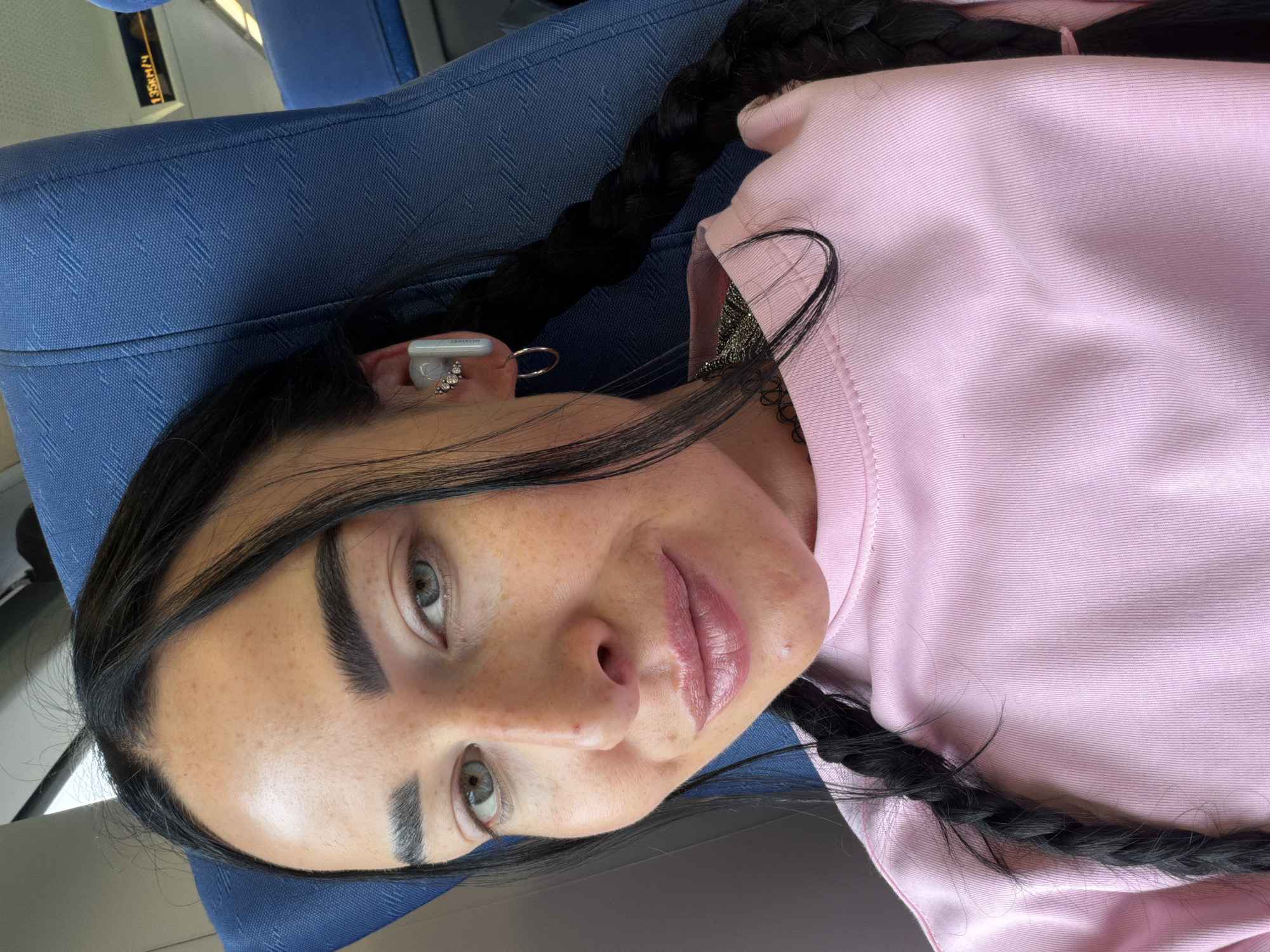The media file shows a close-up, vertically oriented shot of a woman with dark hair in a braid, wearing a pink sweatshirt. She has light-colored eyes, prominent eyebrows, and visible freckles on her face. She is wearing a white earbud in her right ear, which has the branding "HUAWEI" visible. She also has a small, sparkly earring in her ear. Her lips appear to be glossy.
The woman is seated on a blue fabric surface, which appears to be a seat with a patterned texture. In the background, a yellow sign with black text is visible, reading "135km/4". This suggests a transportation setting, possibly a train or bus. The lighting indicates it is daytime. The overall mood is casual and relaxed.
The location context provided is Ушаки, Russia.