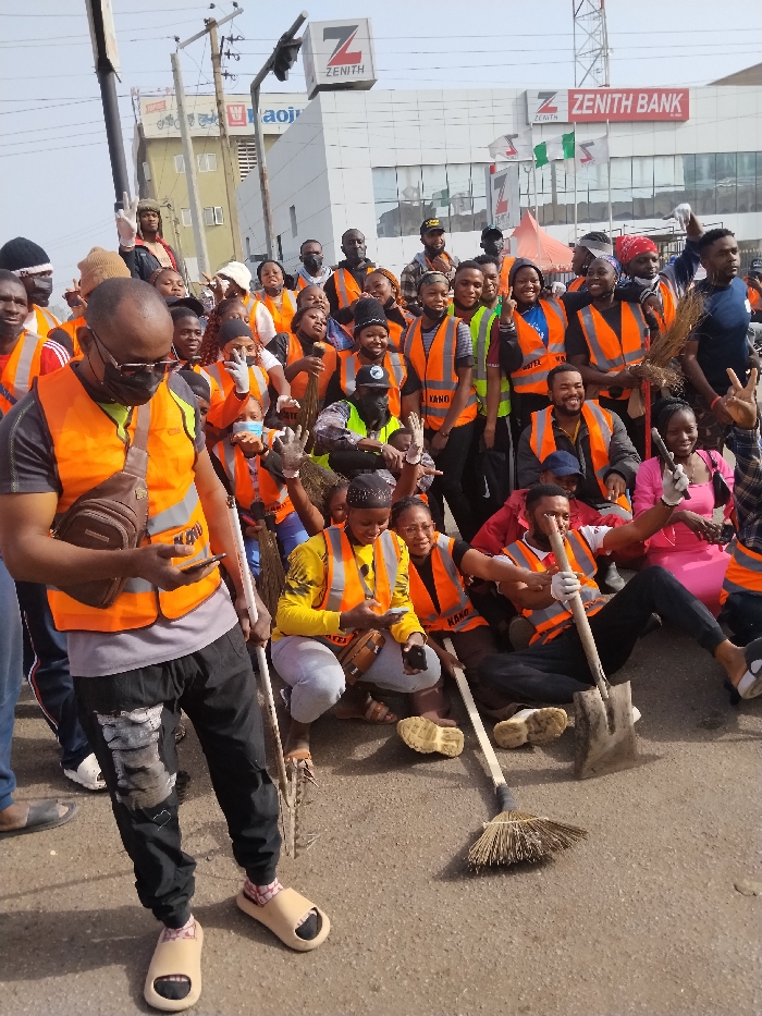 The image depicts a large group of approximately 30-40 individuals, primarily young adults, gathered on a paved urban street in Fagge C, Nigeria. The background features a multi-story white building prominently displaying the "Zenith Bank" logo and signage, including a large 'Z' and 'ZENITH' text, along with Nigerian flags. Another building with "aoju" visible text is present further down the street, beneath overhead power lines and a traffic signal structure.
Most individuals in the group are wearing bright orange reflective safety vests, some with "KANO" text. Many are holding cleaning tools, specifically traditional brooms made of bundled sticks. One man seated in the lower right holds a shovel. Several people are wearing white gloves. The group is largely clustered, posing for the camera with various expressions, including smiles, peace signs, and thumbs-up gestures.
In the foreground left, a man wearing a black t-shirt, orange vest, dark sunglasses, and light-colored slip-on sandals stands, holding a white-handled broom and looking down at a smartphone in his right hand. A brown sling bag is worn across his chest. Two women crouching in the mid-ground also appear to be looking at their phones. The ground is a light gray asphalt surface, and the sky is bright and clear, indicating daylight.