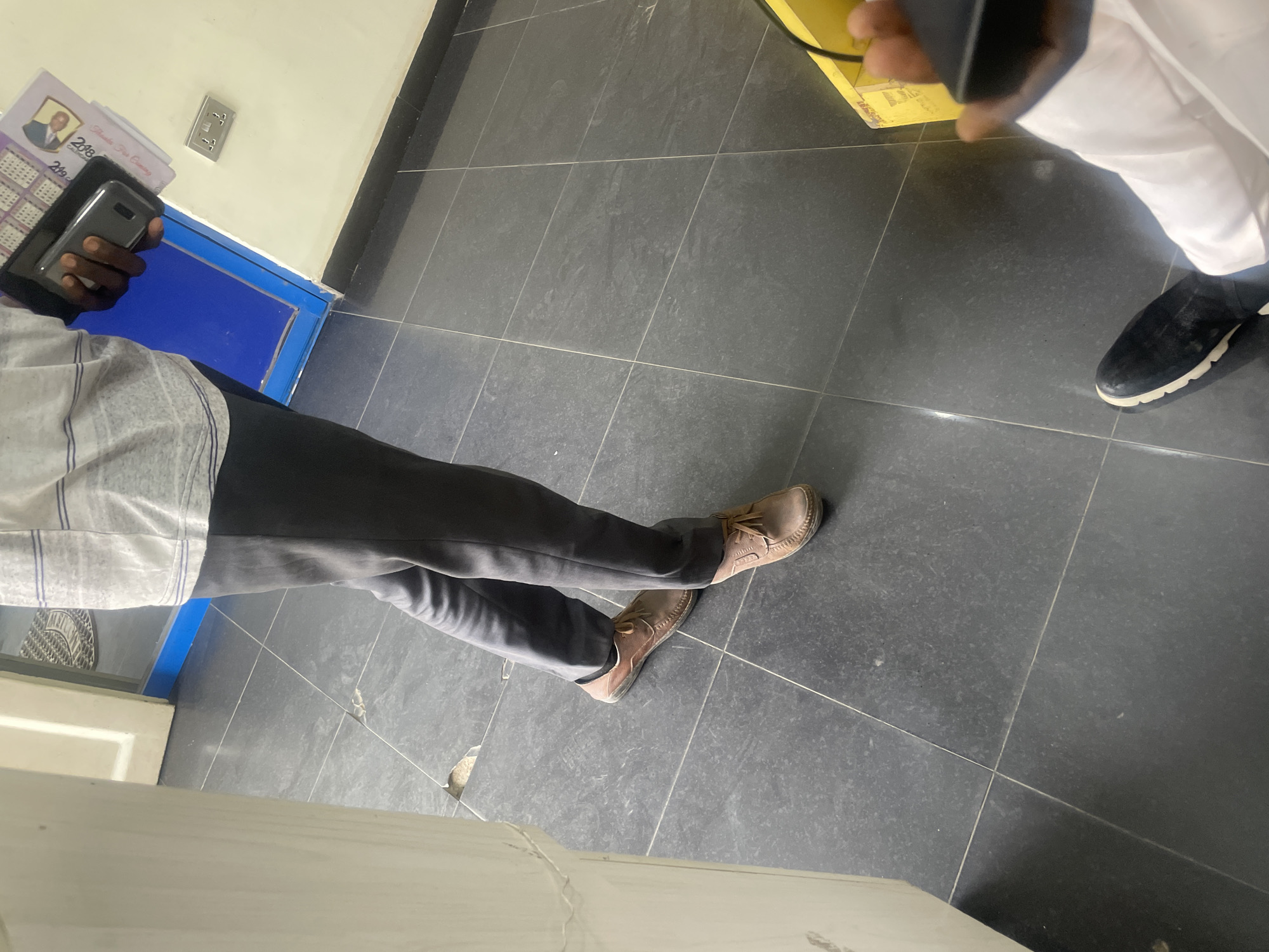 The image shows the lower body and hands of two people standing on a tiled floor. One person, wearing dark pants and brown shoes, is holding a cell phone and a calendar or booklet. The other person, wearing white clothing and black shoes, is partially visible in the foreground. The environment appears to be indoors, possibly a commercial space, with a blue-framed door and a light-colored wall with electrical outlets visible in the background. The scene is set in Ikeja, Nigeria.