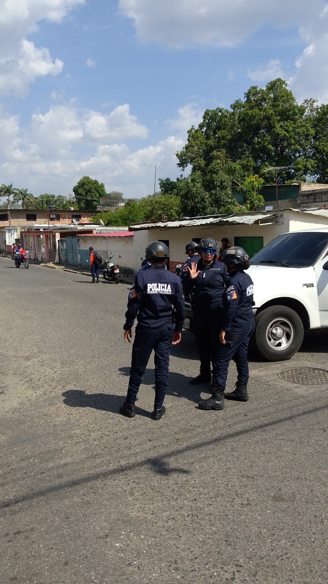 The image shows a street scene in Tinaquillo, Venezuela. Three uniformed police officers stand in the middle of the road, near a white Ford F-150 pickup truck. The officer in the center is facing the camera with their left hand raised. The officers are wearing helmets, dark blue uniforms with "POLICIA" and "COJEDES VENEZUELA" printed on the back. Two motorcyclists are on the left side of the road, while another pedestrian is standing near the sidewalk. Buildings and trees are visible in the background under a partly cloudy sky.