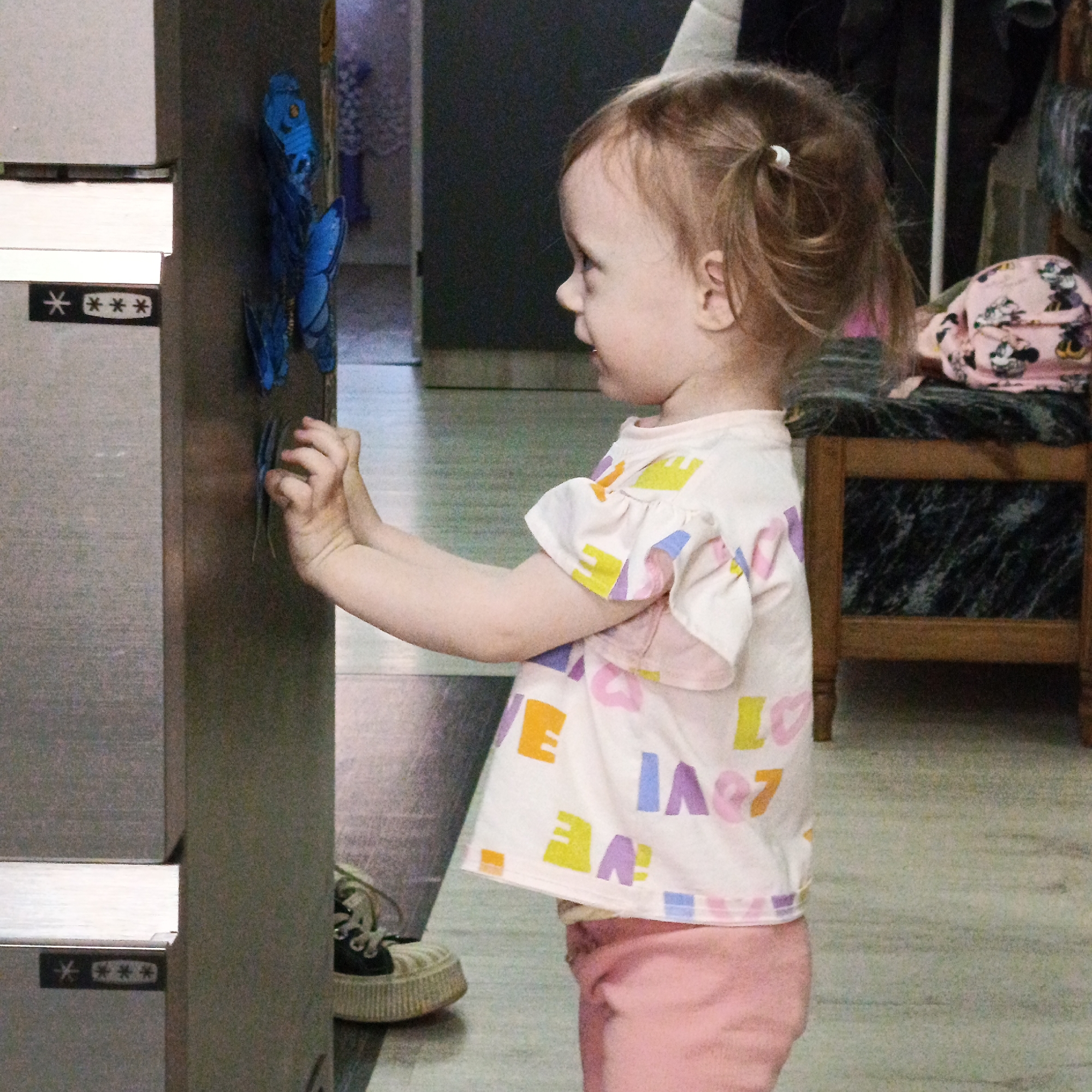 A young child is standing in front of a gray metal cabinet, possibly a refrigerator, with two blue plastic butterflies attached to the surface. The child has light skin, brown hair pulled back in two small ponytails, and is wearing a white t-shirt with colorful geometric shapes and the word "LOVE" printed on it, along with pink pants. They are touching one of the butterflies with both hands. The background includes a door, a wooden chair with a pink patterned cushion, and a pair of sneakers on the floor near the cabinet. The location is possibly in the city of Марковский, Russia, as the prompt suggests, though there is no concrete evidence within the image to confirm this.